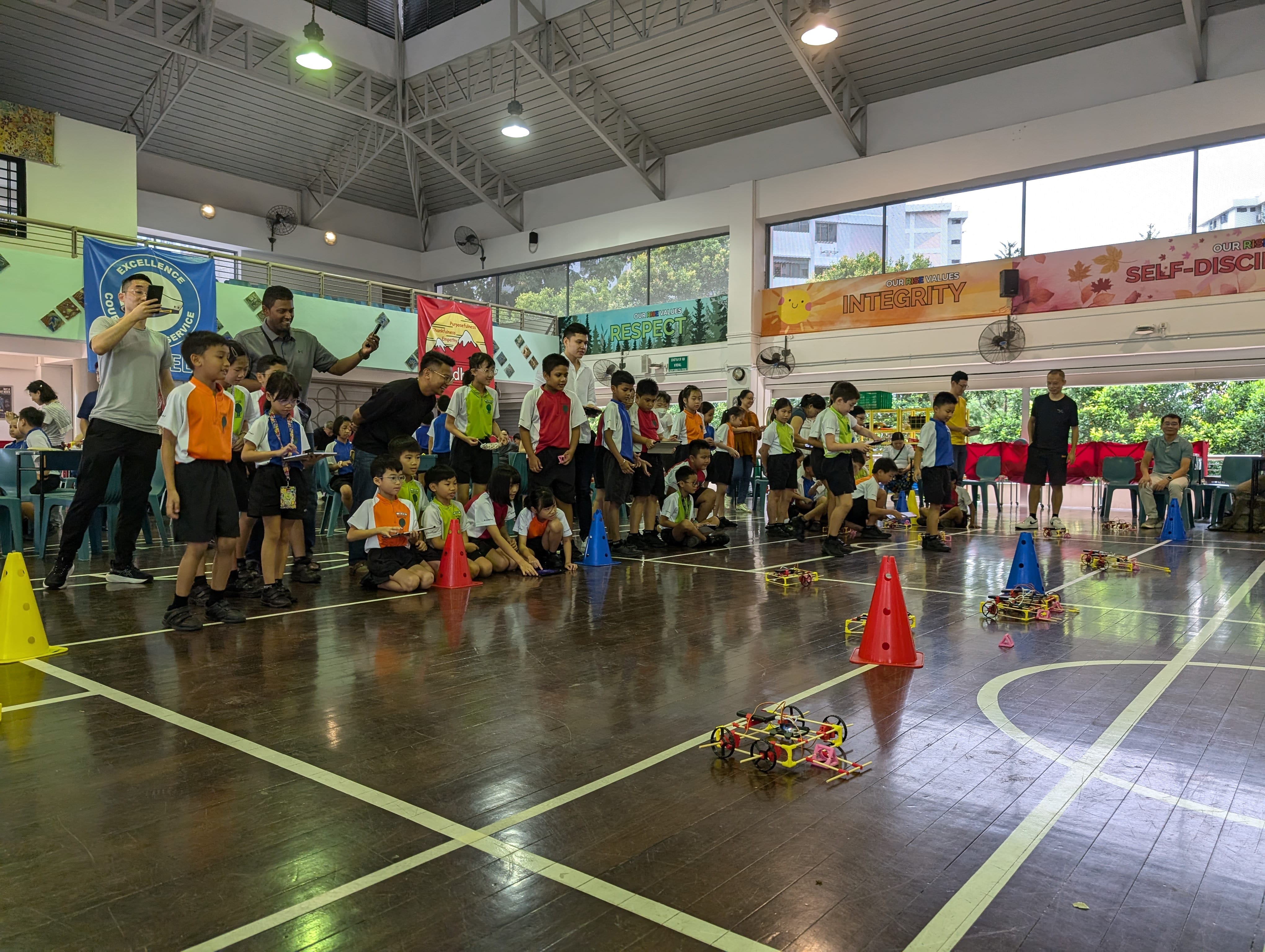 Students in a gym watch small robots race between colored cones; teachers stand nearby filming.