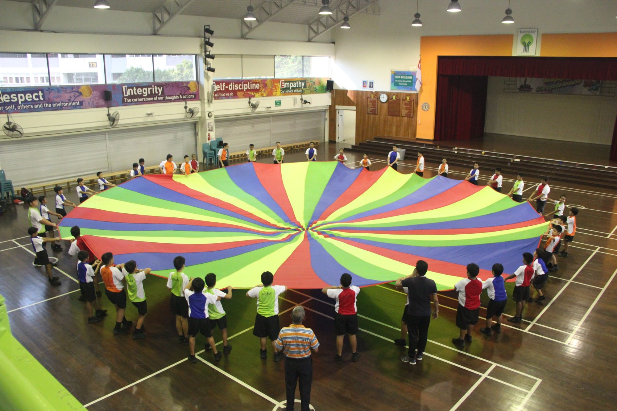 Students stand in a circle holding a large colorful rainbow parachute in a school gymnasium.