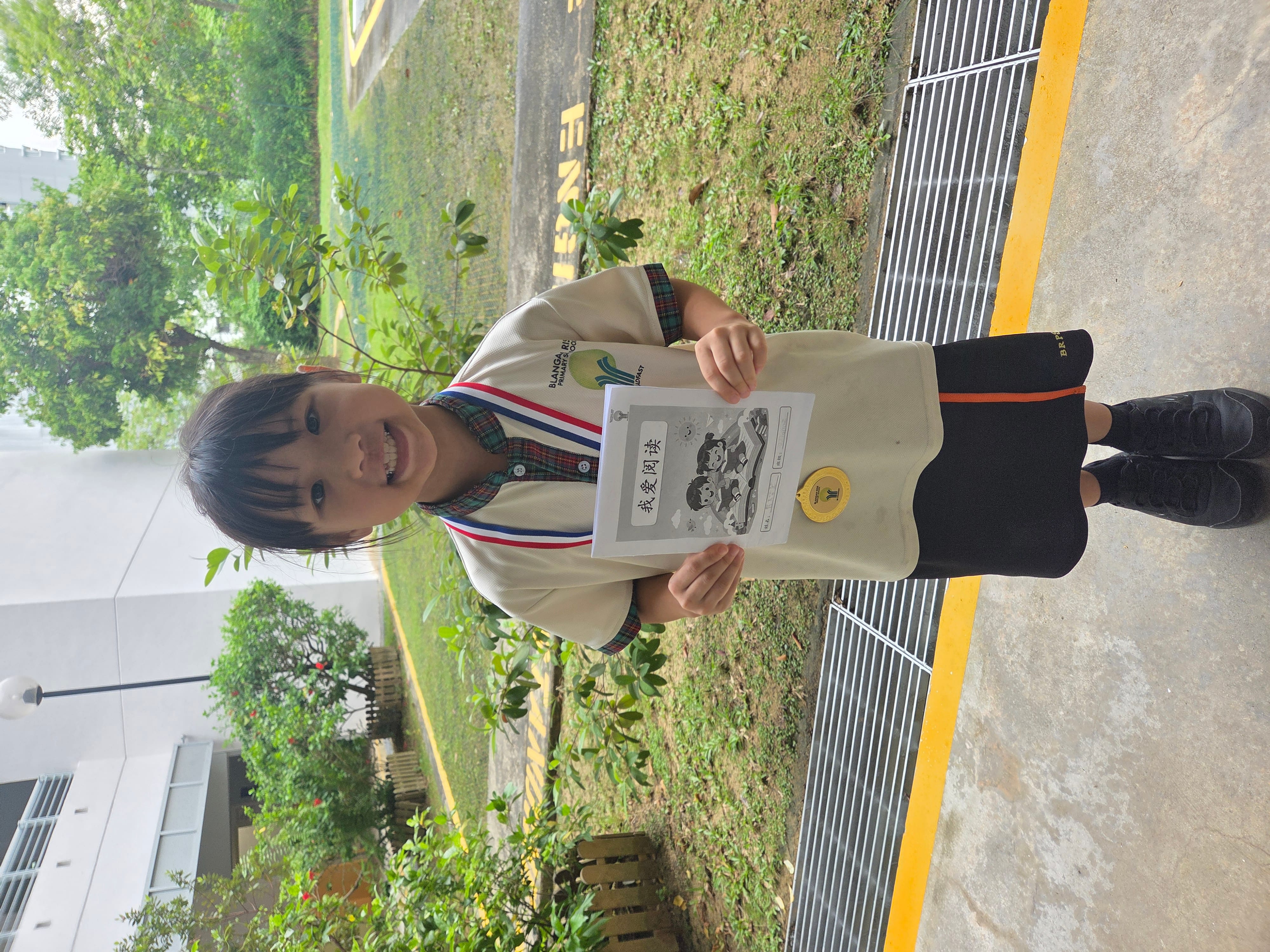 Smiling child wearing a medal holds a certificate while standing on a wet sidewalk by greenery.