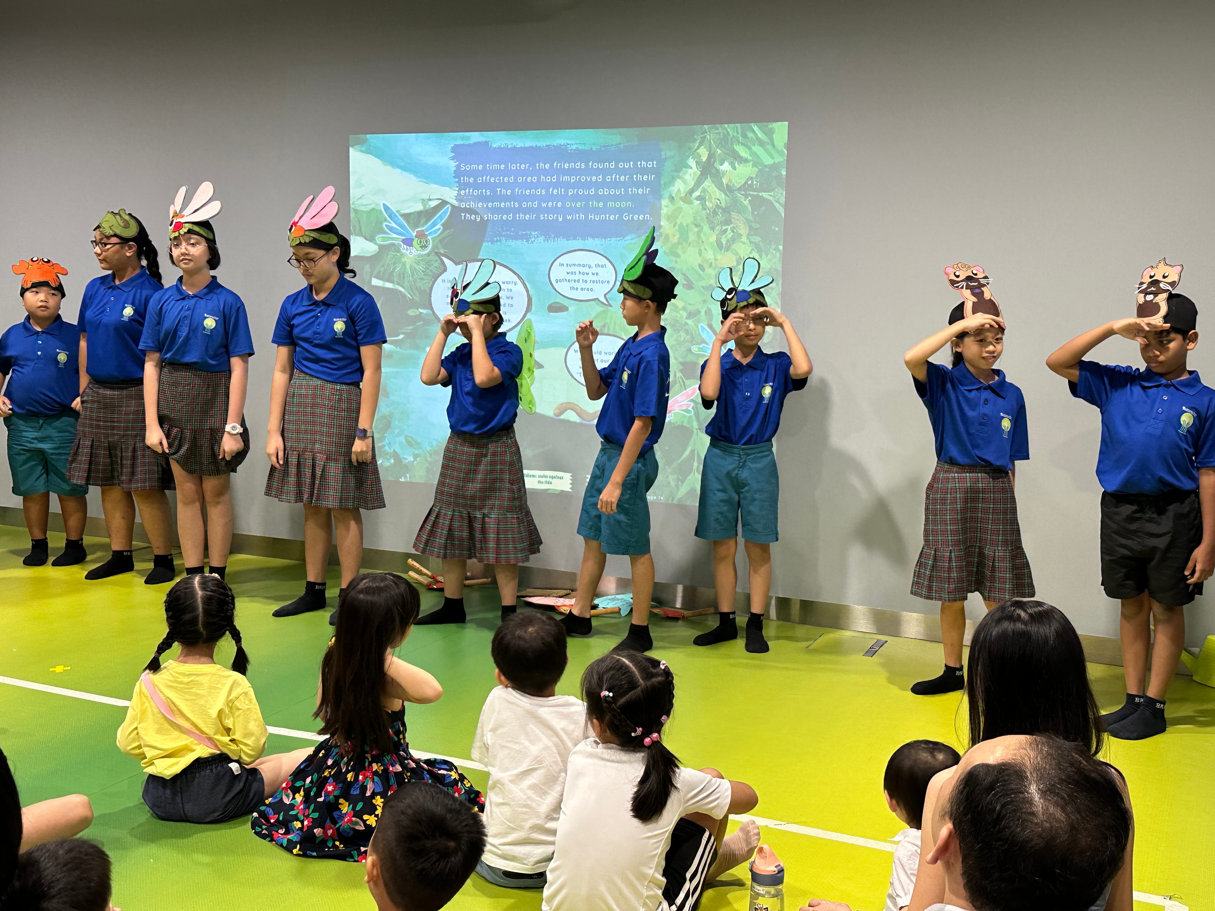 Children in blue uniforms perform in front of a projected scene, wearing animal headbands; audience sits on floor.