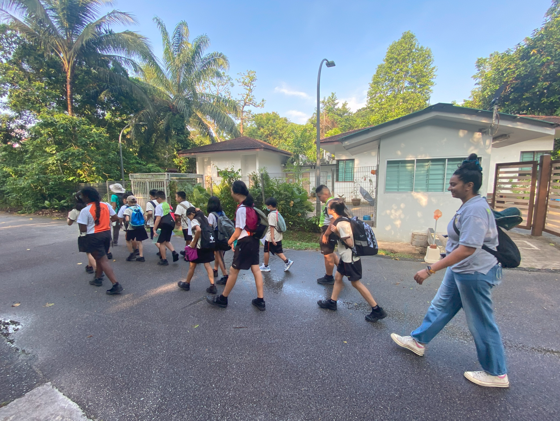 Group of schoolchildren with backpacks walk along a road past houses and tall palm trees, with an adult nearby.