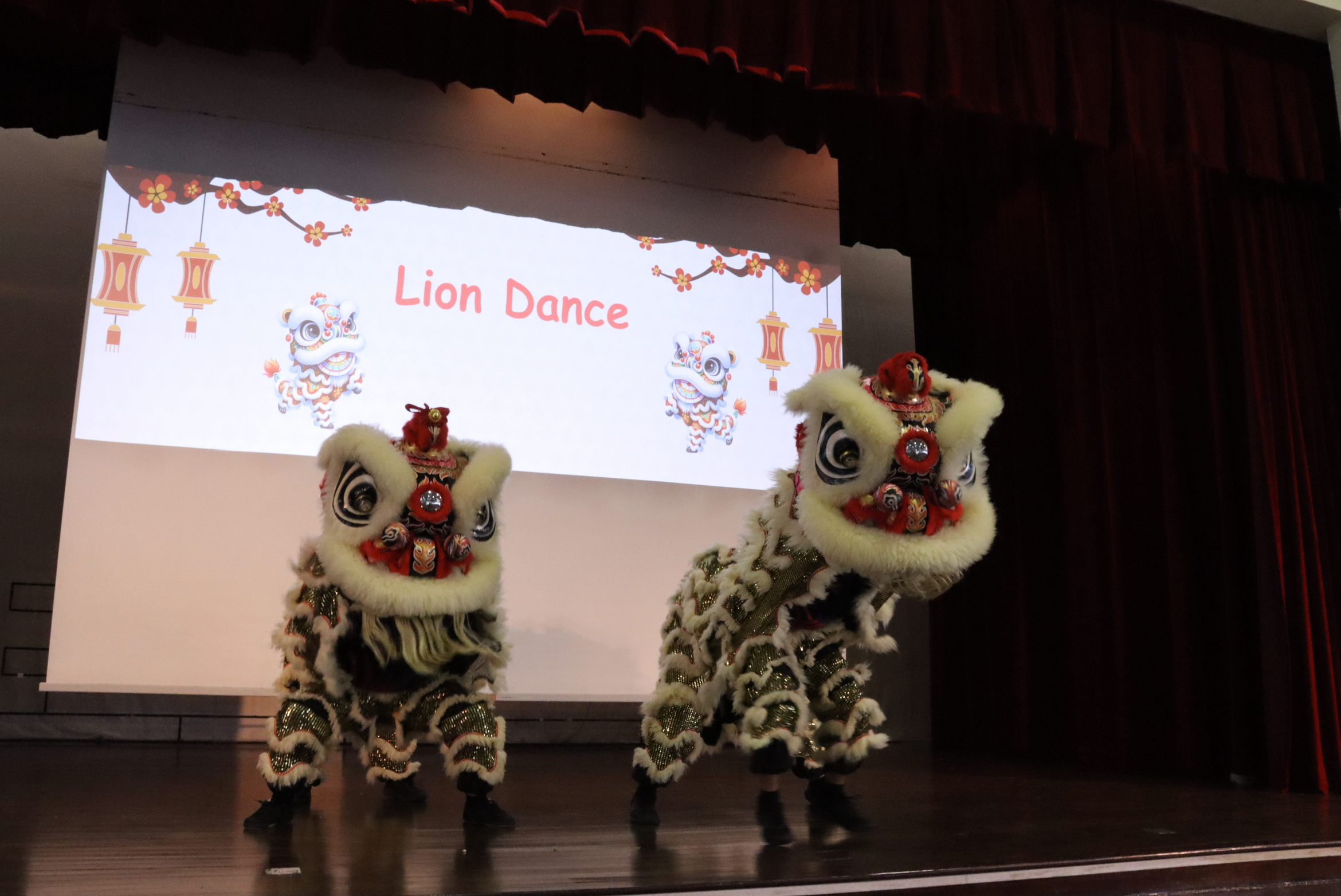 Two lion dance costumes perform on stage in front of a screen reading Lion Dance with lantern graphics.