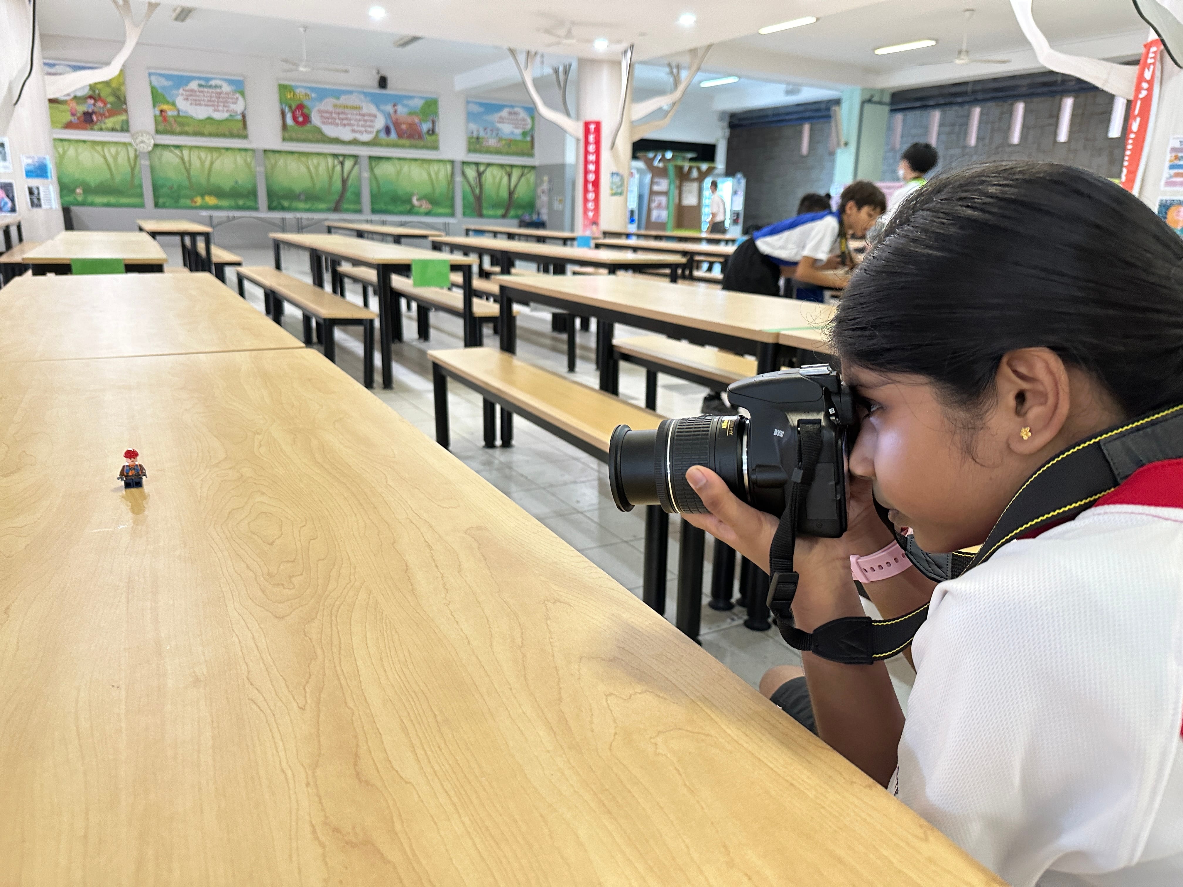 A girl photographs a small Lego minifigure on a wooden table in a school cafeteria with colorful murals.