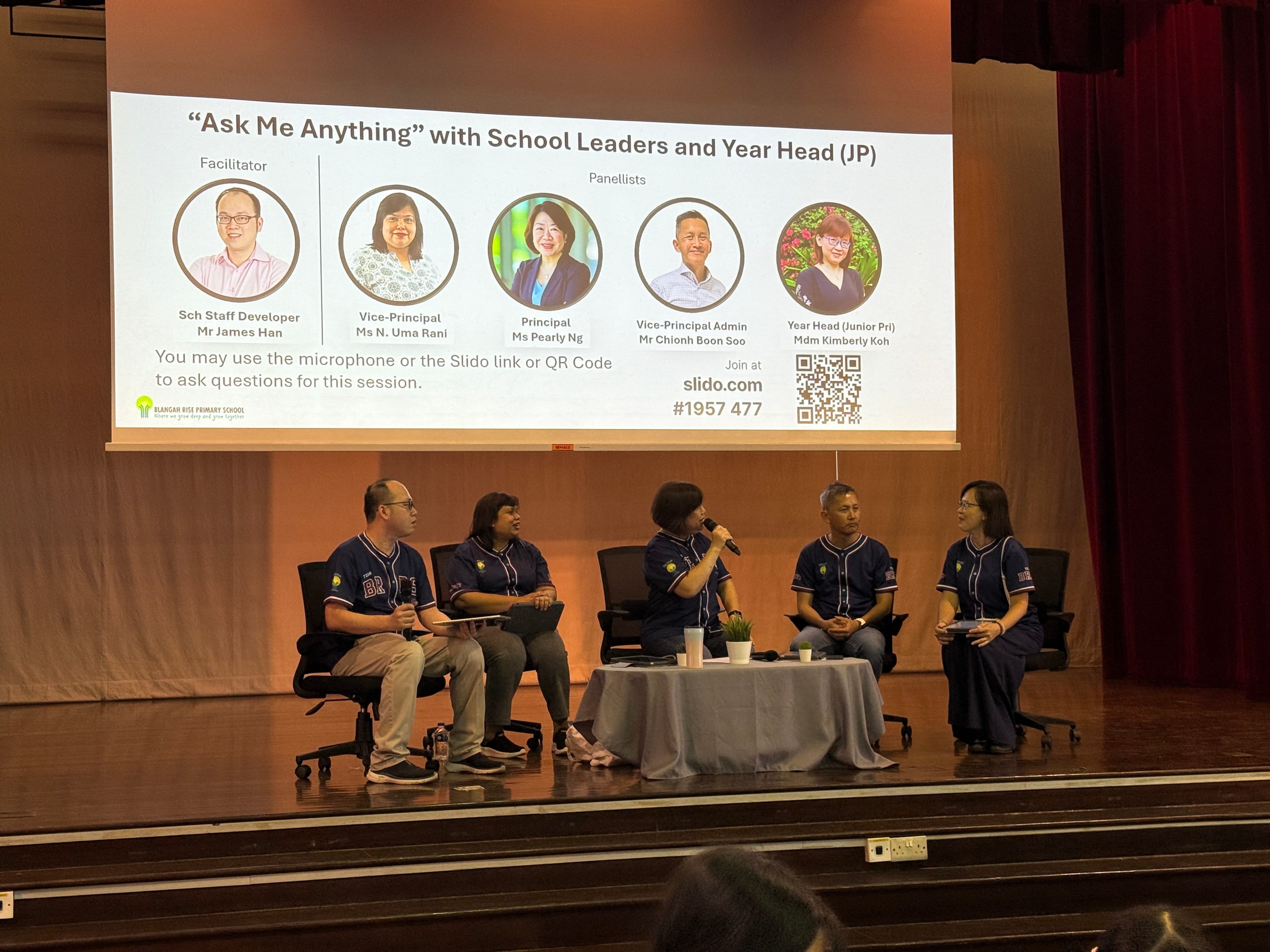 Five panelists in navy shirts sit on stage during an Ask Me Anything session with school leaders shown on a projected slide.