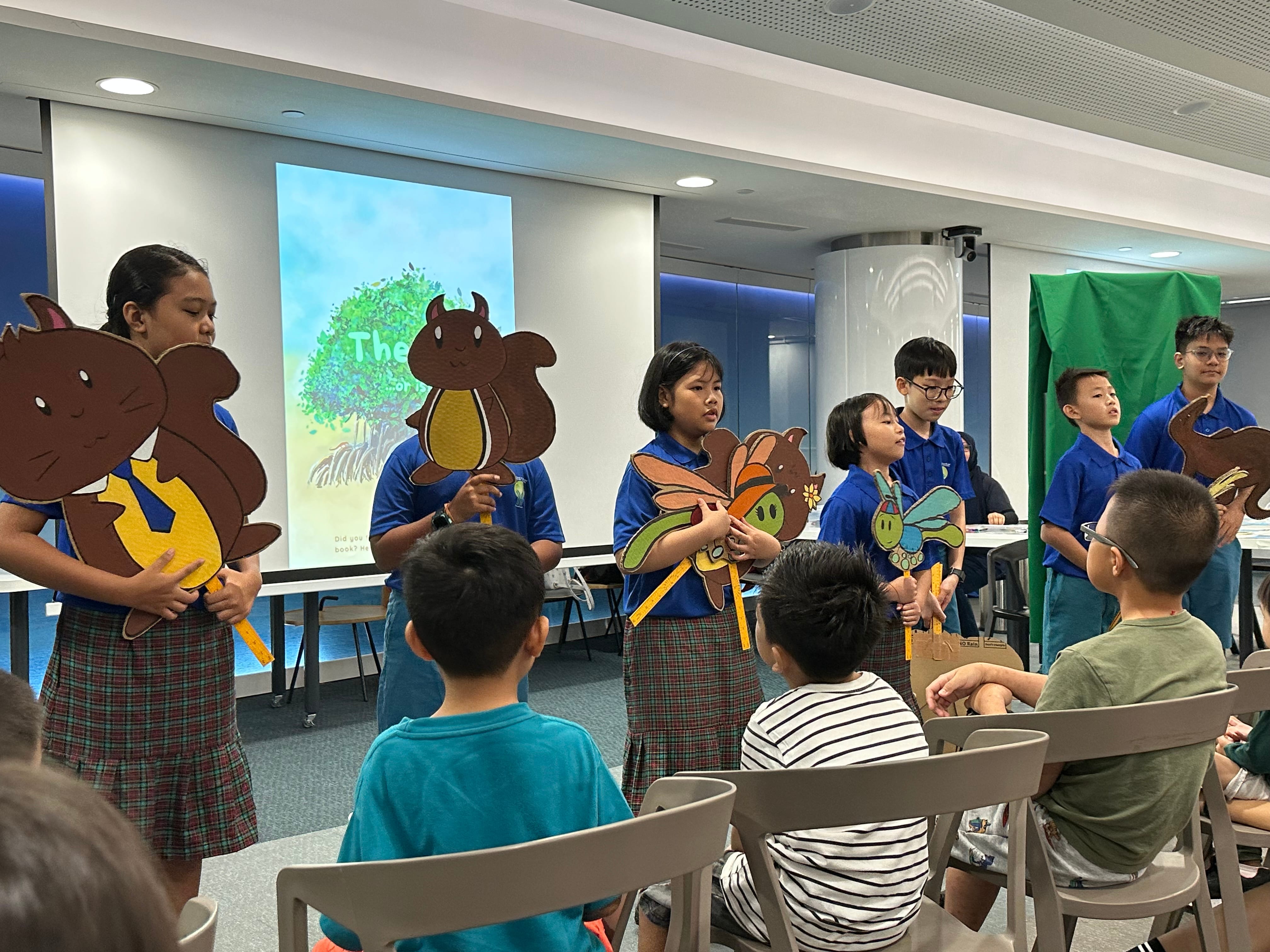 Children in blue uniforms hold animal cutouts on sticks, performing a story for young seated kids in a bright room.