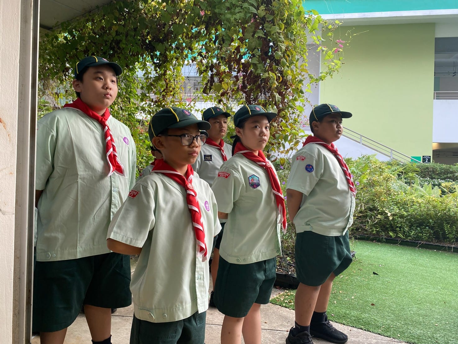 Five children in scout uniforms with green caps and red neckerchiefs stand attentively outdoors near a building.