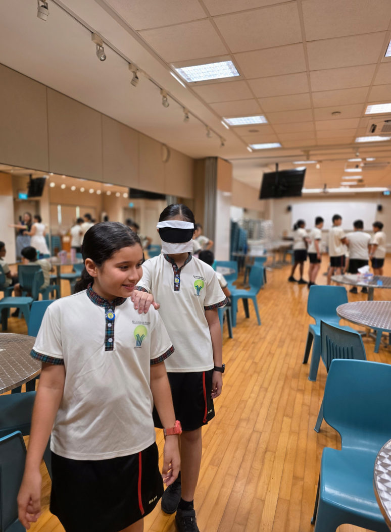 Blindfolded student follows another with a hand on their shoulder in a room with tables, chairs, and groups.