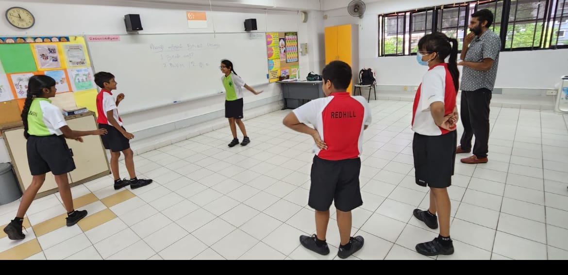 Several students in Redhill school uniforms stand in a classroom while a teacher observes near a whiteboard.