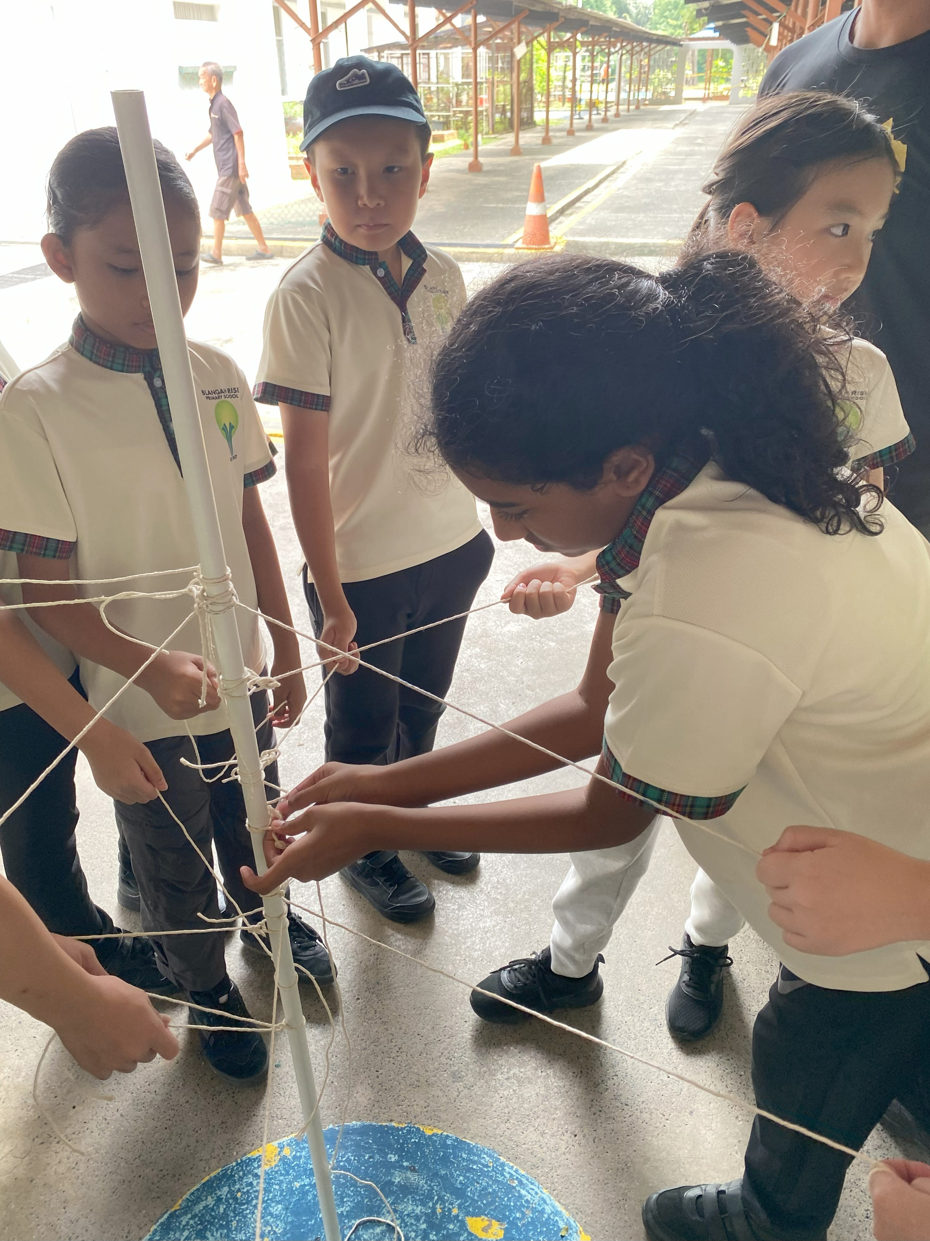 Several primary school students in uniforms work together tying ropes around a white pole outdoors.