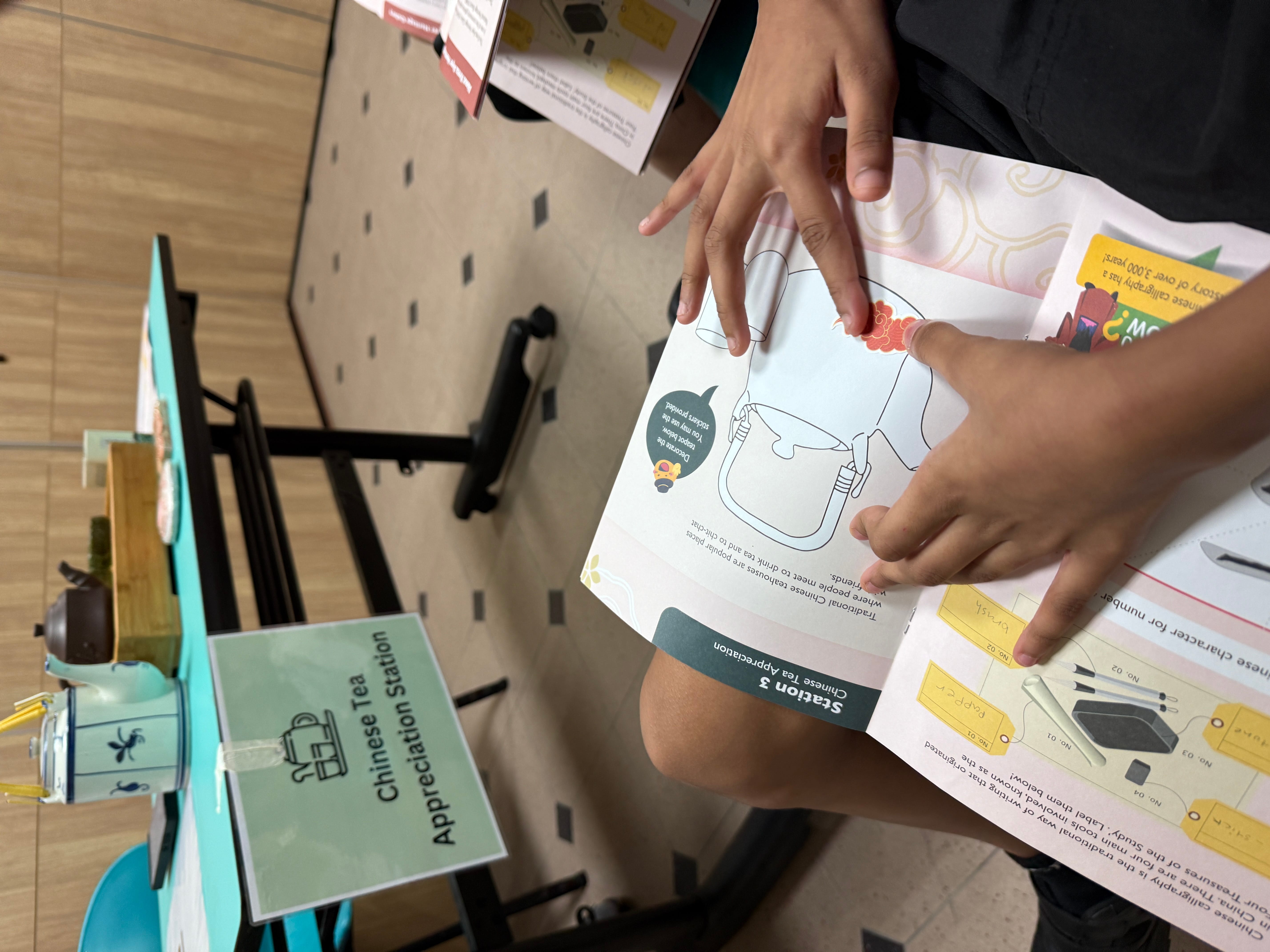 Child's hands holding a booklet open to Station 3 Chinese Tea Appreciation, with a tea station and teapots in the background.