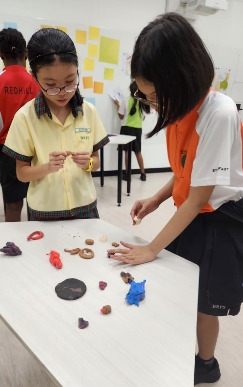 Two school girls in uniforms work with colorful clay sculptures on a white table in a classroom.