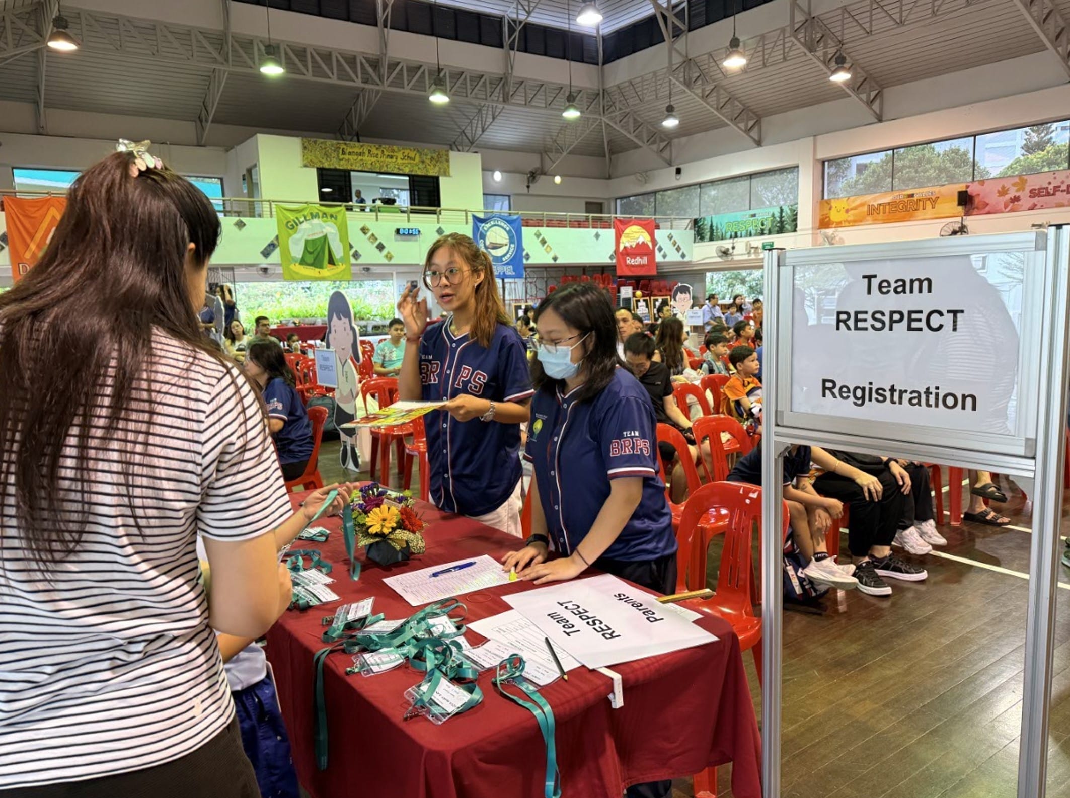 Two staff in blue BRPS jerseys assist a visitor at a Team RESPECT Registration table in a school hall.