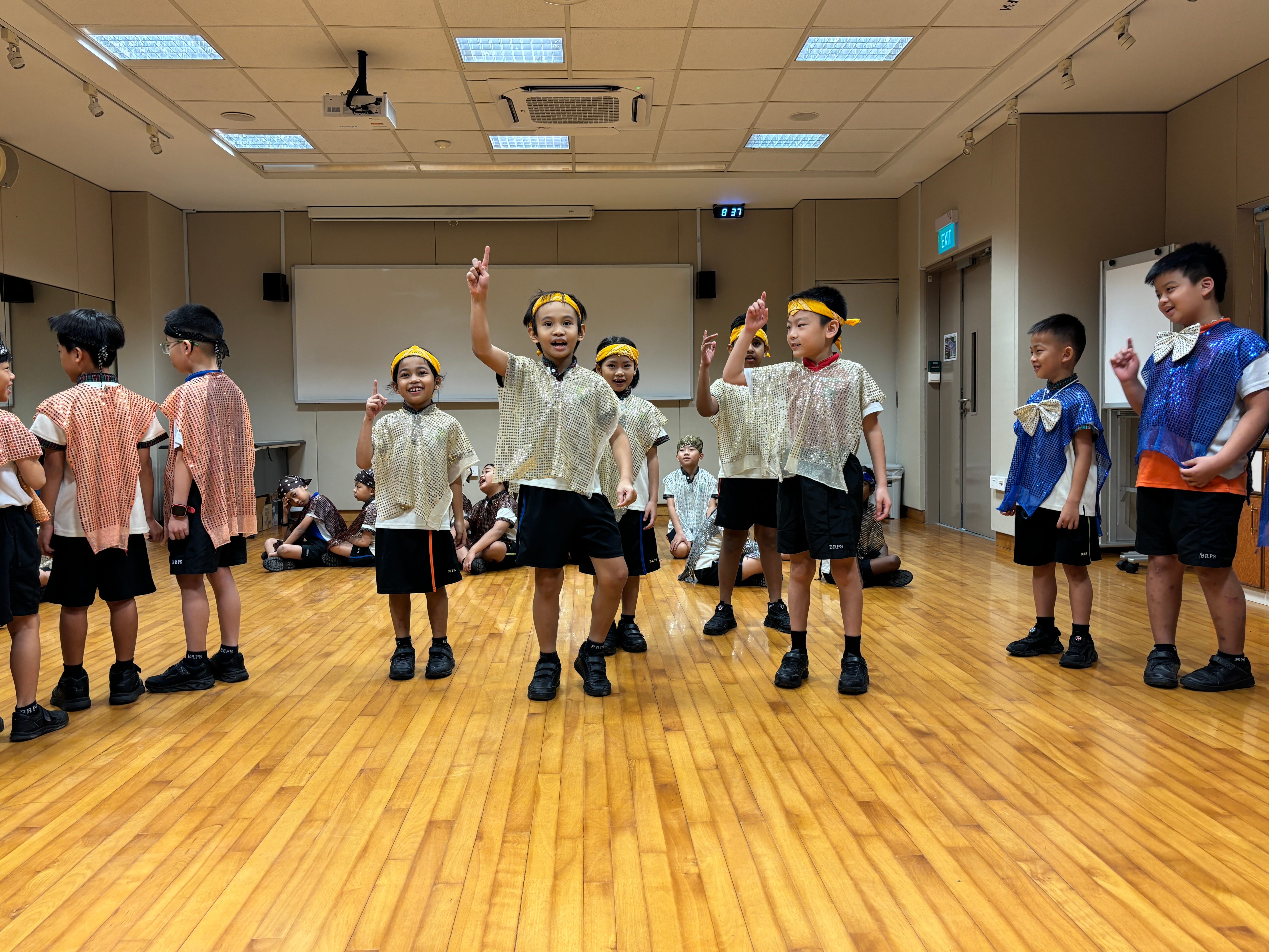 Group of boys in shiny costumes and headbands perform on a wooden floor, some pointing upward.