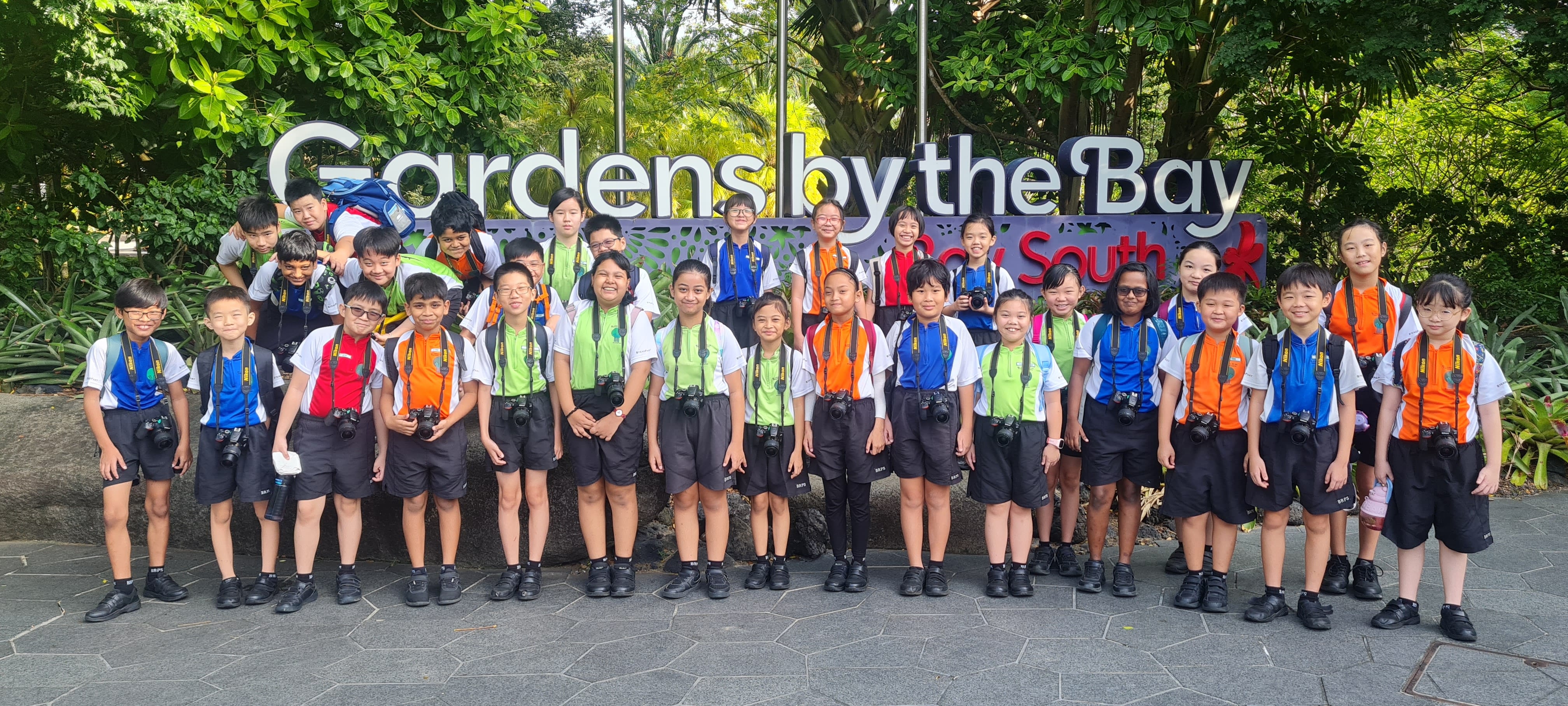 A group of children wearing cameras pose in front of the Gardens by the Bay sign surrounded by green trees.