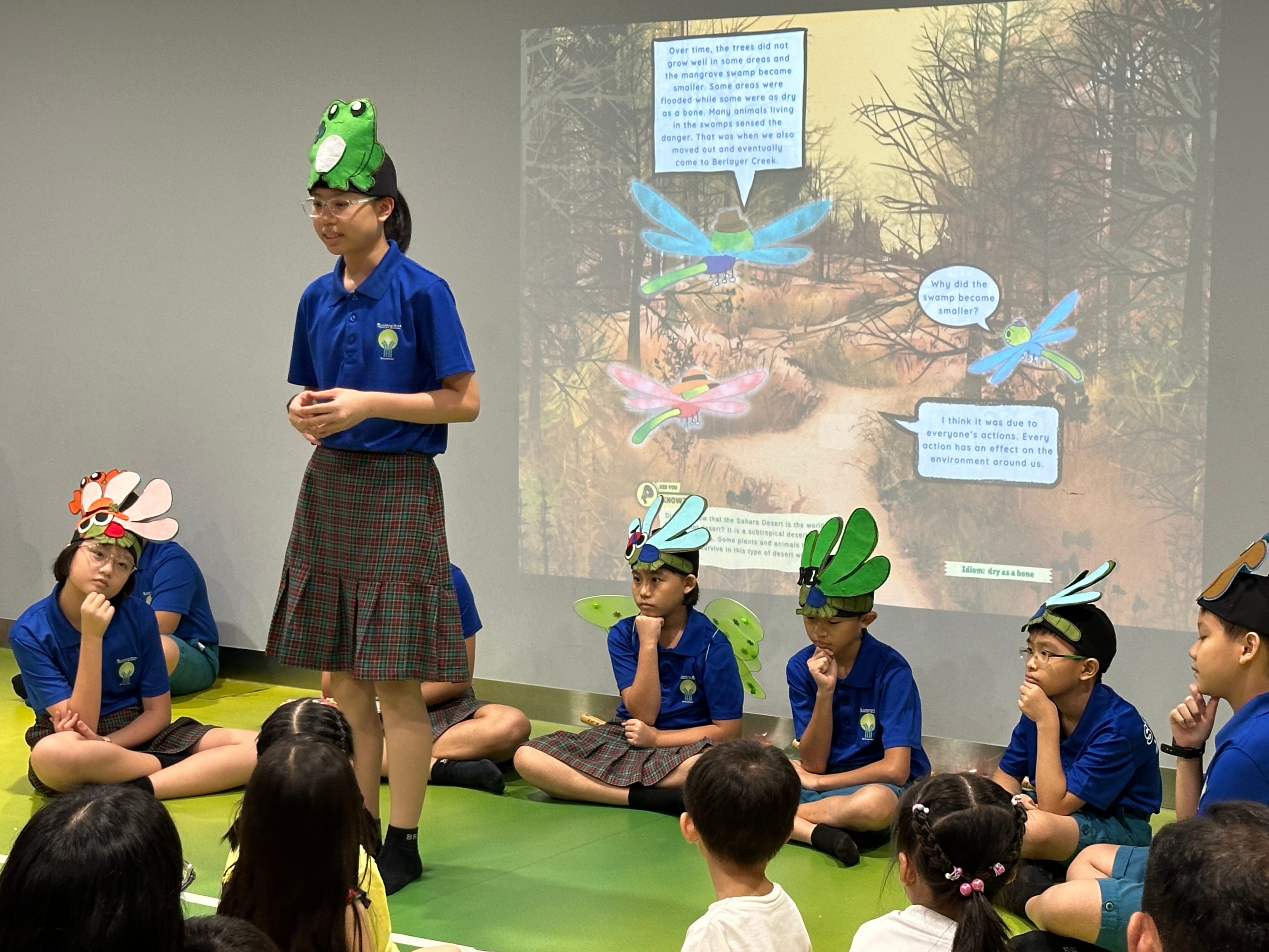 Student in frog hat presents to seated classmates in insect hats, with a projected storyboard behind them.