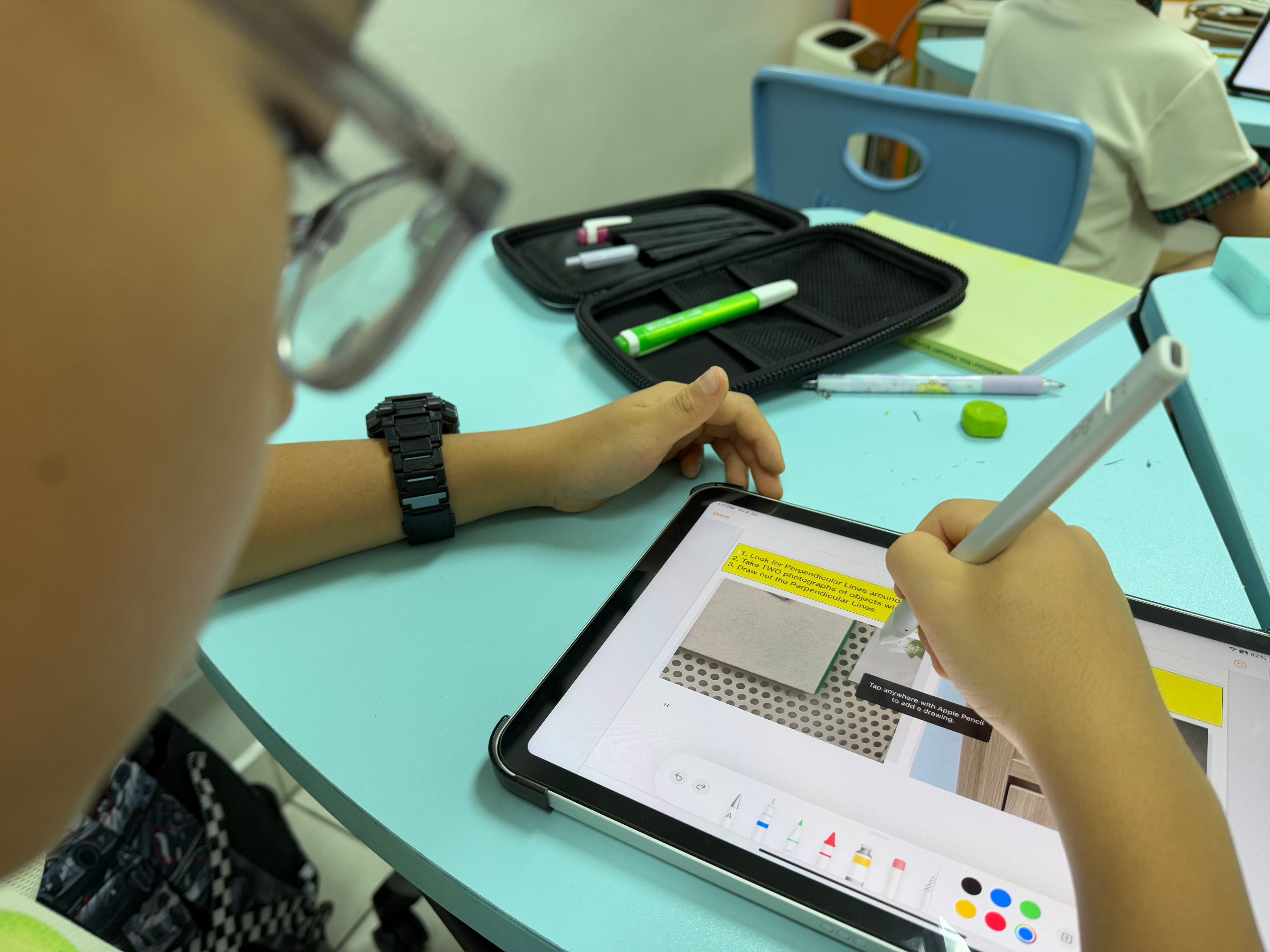 Student wearing glasses uses a stylus on a tablet at a desk with a pencil case and stationery.