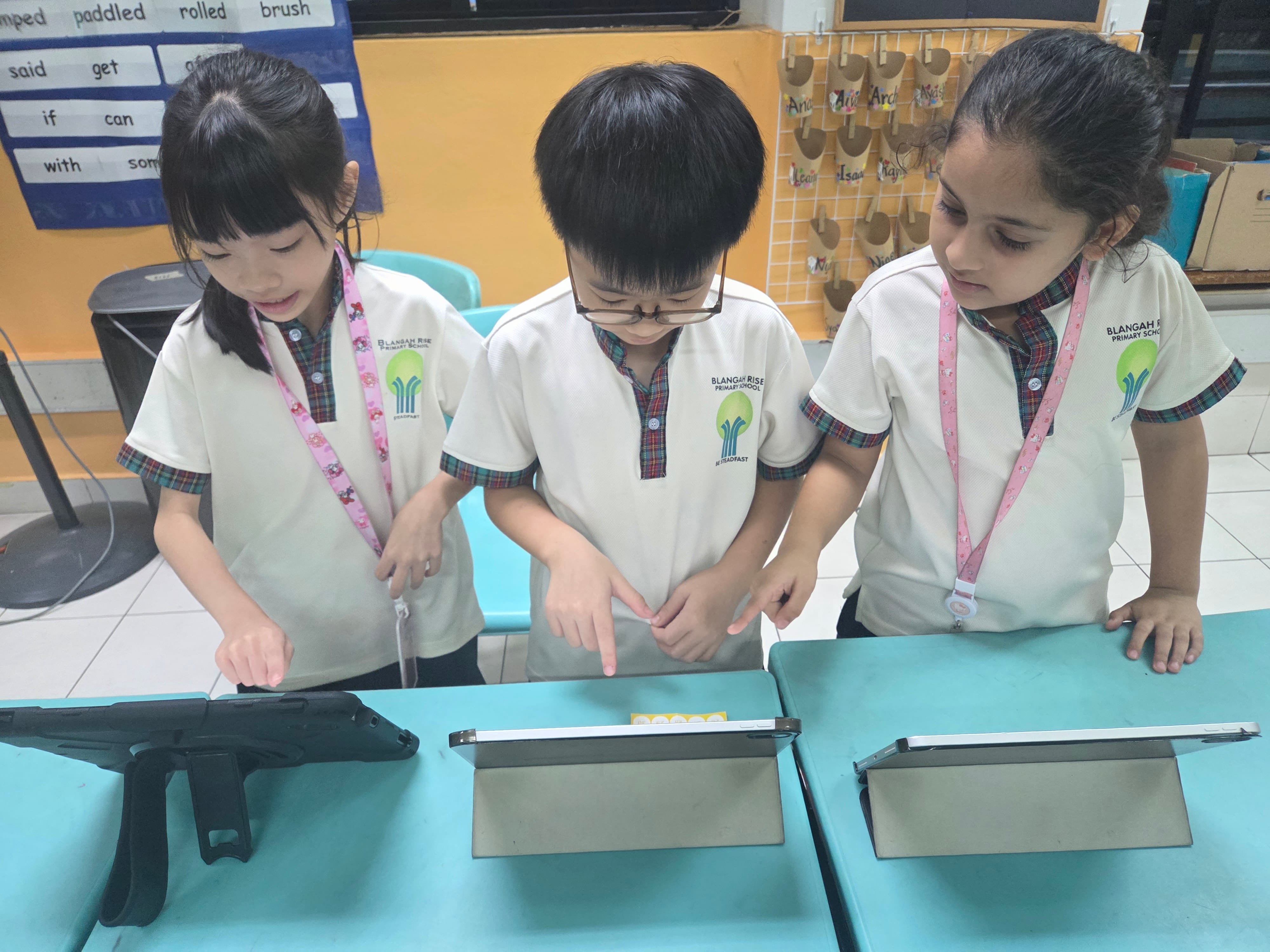 Three schoolchildren in uniforms stand at a table, looking down and pointing at tablets.