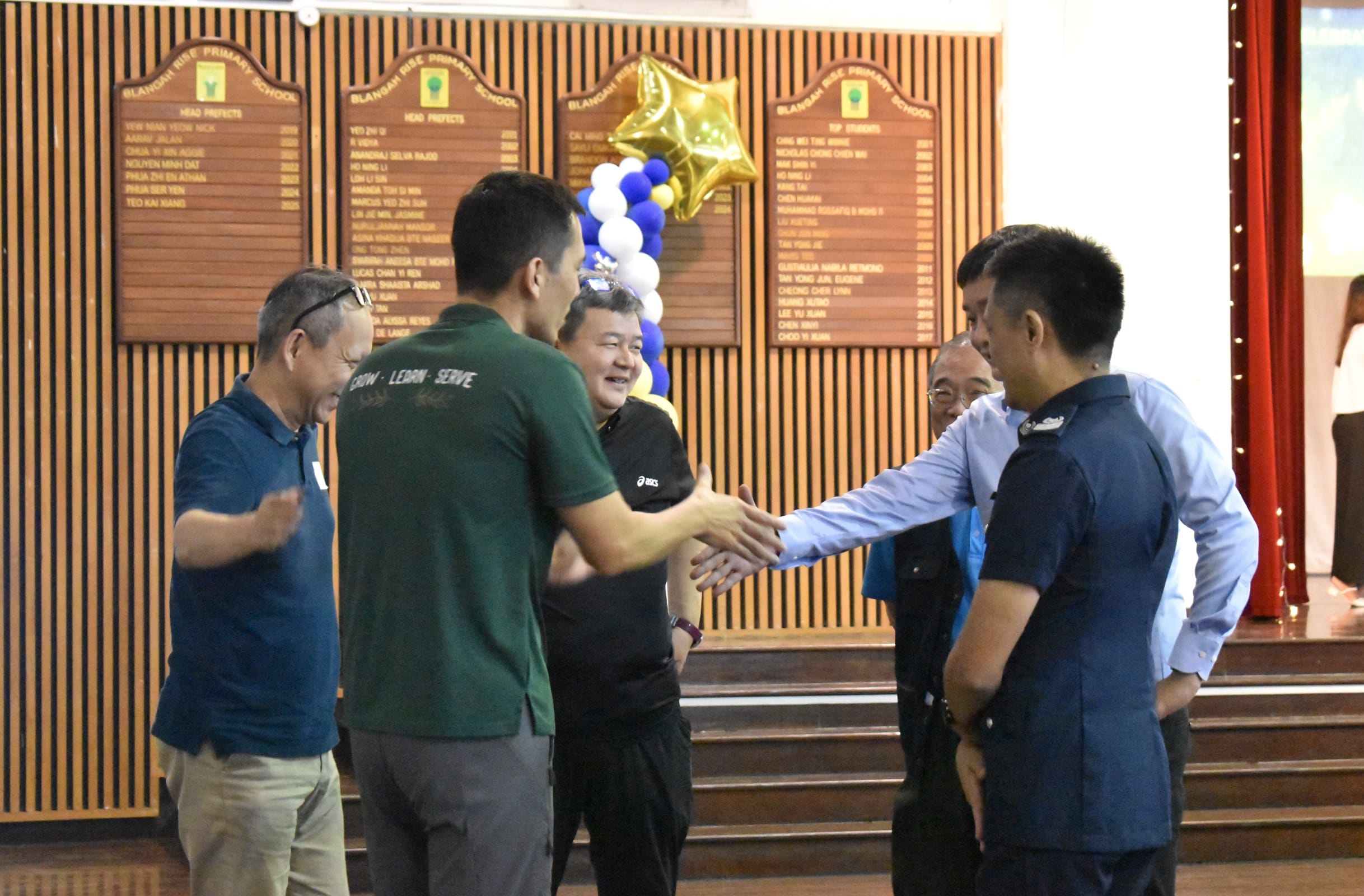 Several men shaking hands and smiling in a school hall with honor boards and blue and gold balloons.