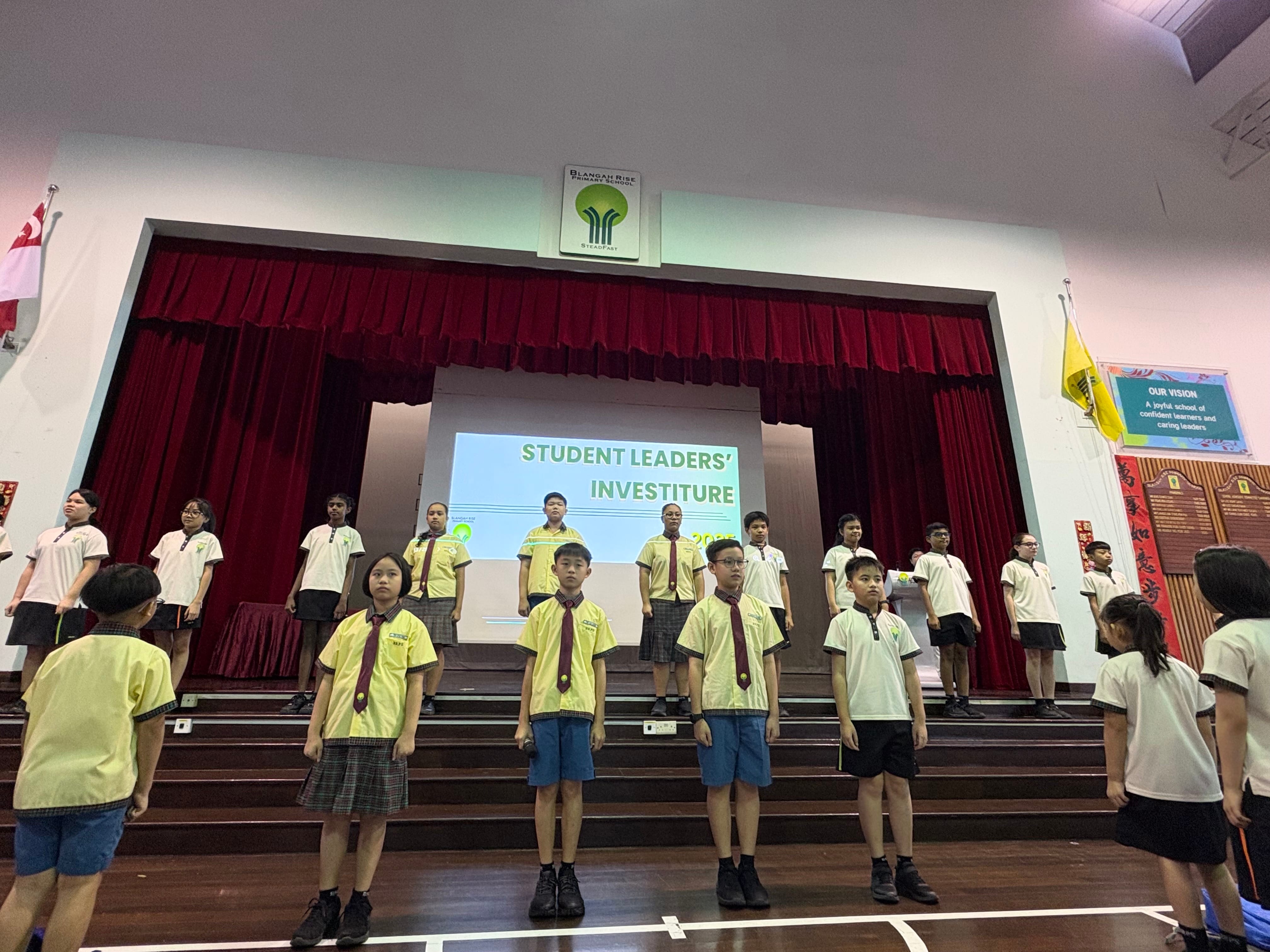 Students in yellow uniforms stand at attention during a Student Leaders Investiture ceremony on a school hall stage.