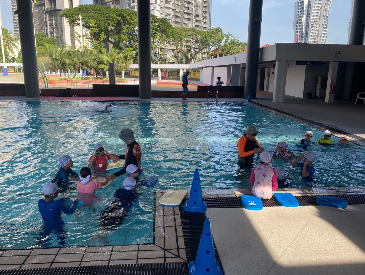 Children in swim caps and goggles receive swimming lessons in an outdoor pool, guided by two instructors.