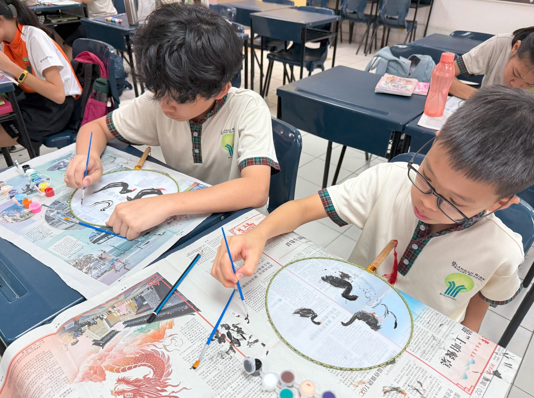 Two school boys paint black dragon designs on circular fans using brushes and ink in a classroom art session.