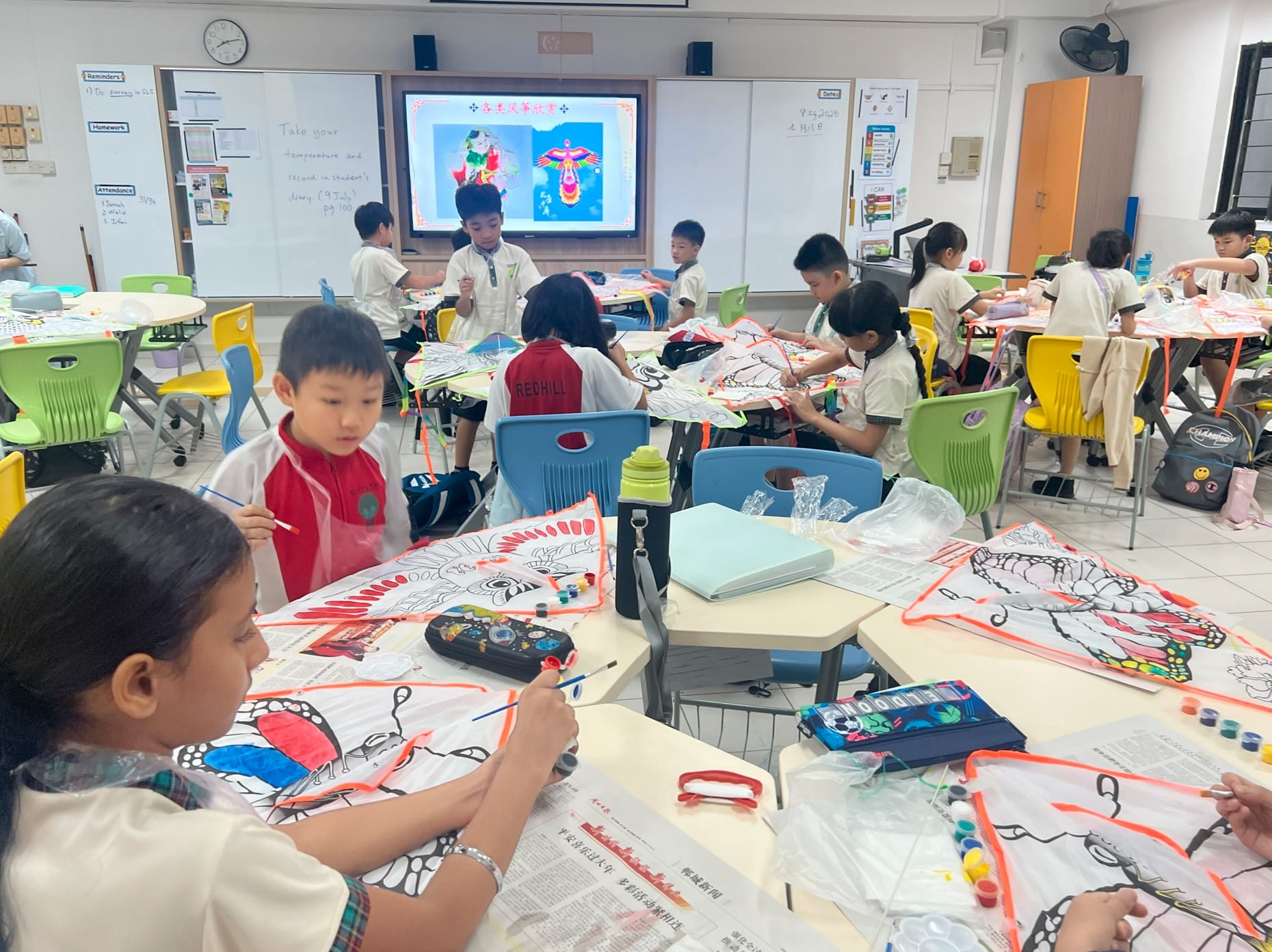 Young students in school uniforms paint kite outlines at group desks in a classroom with a whiteboard and display screen.