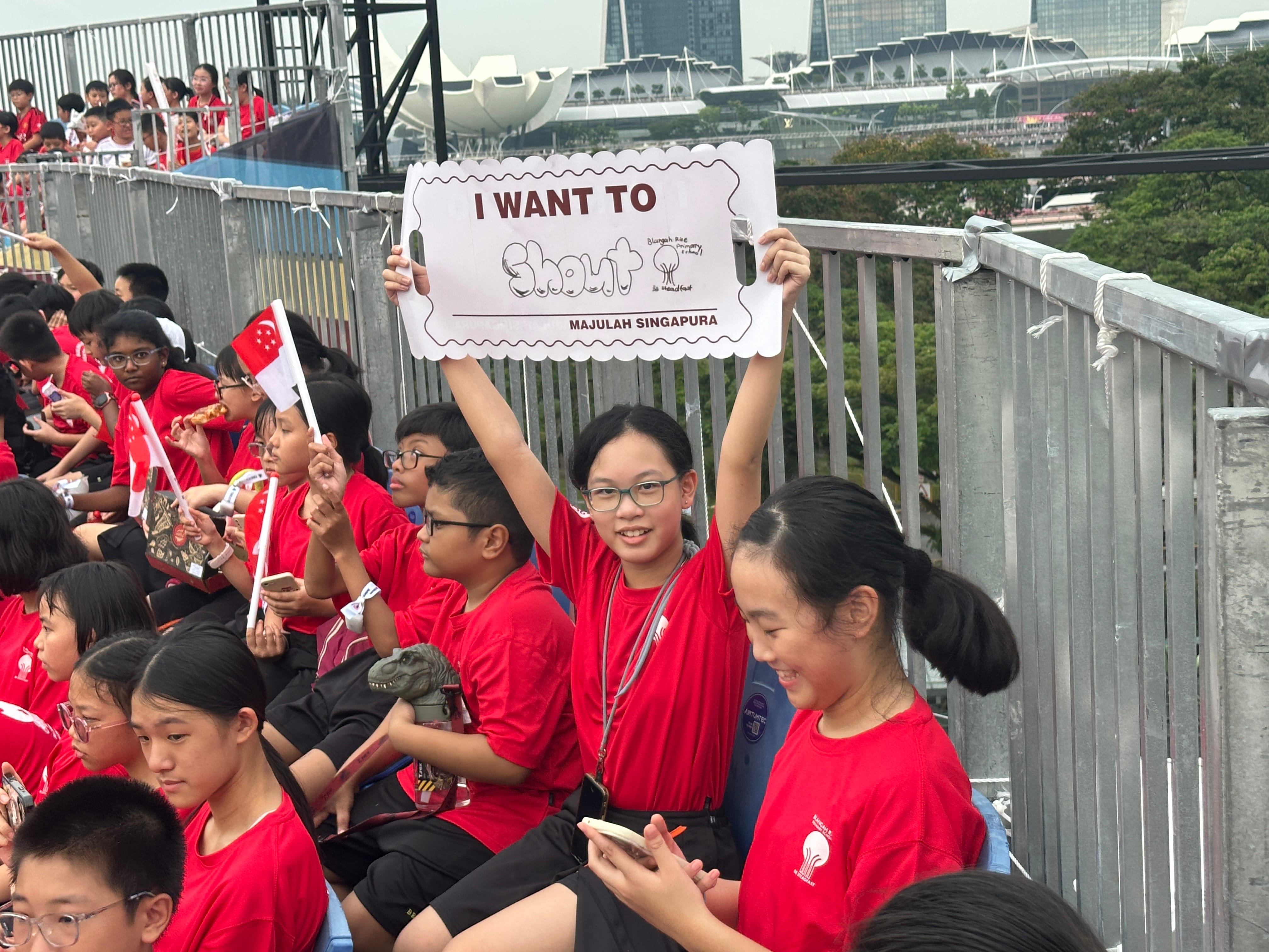 A girl in a red shirt holds a sign reading "I Want To Shout Majulah Singapura" among children waving Singapore flags.