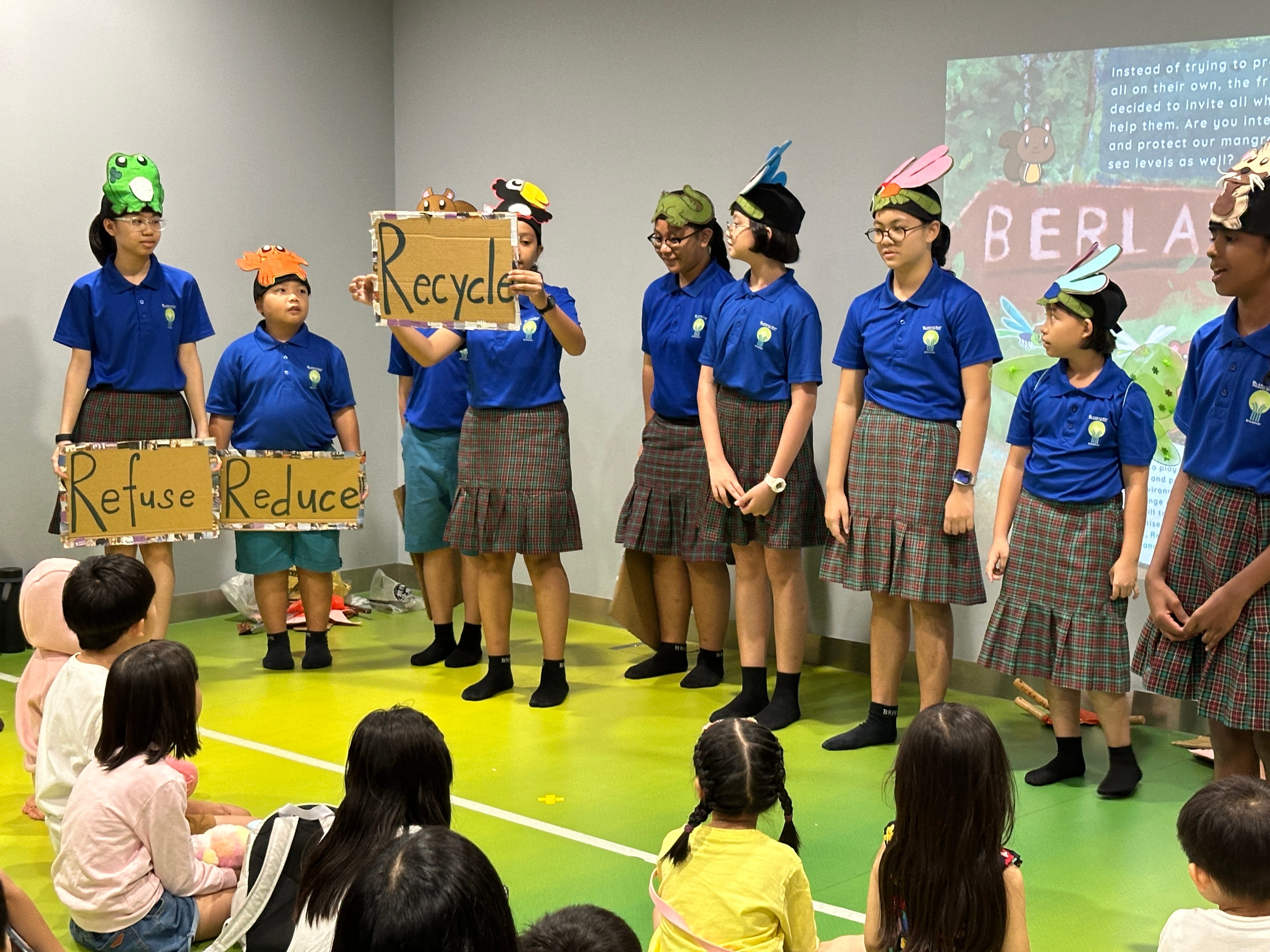 Students in blue uniforms hold cardboard signs reading Refuse, Reduce, and Recycle during an environmental skit for younger children.