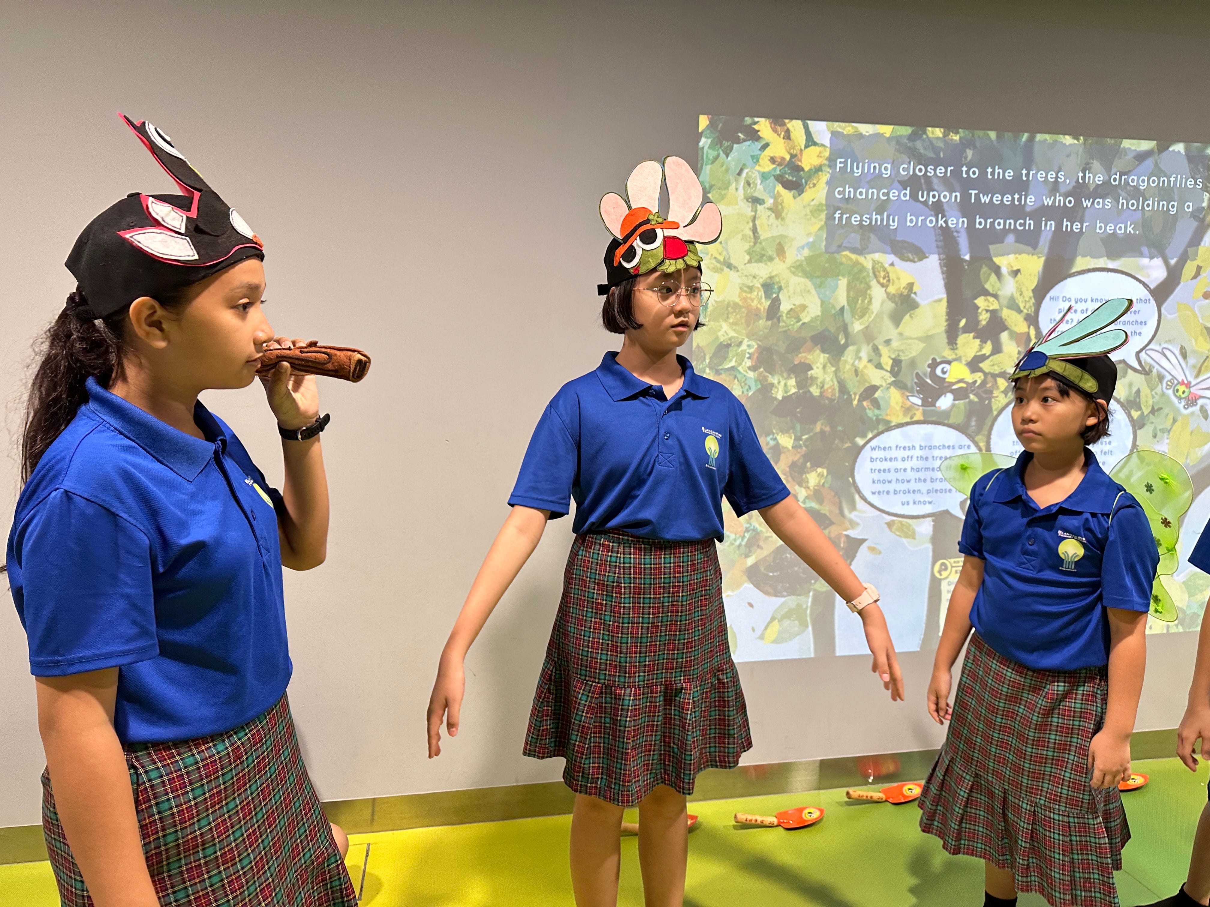 Three children in blue uniforms and plaid skirts wear insect masks, acting in front of a projected story scene.