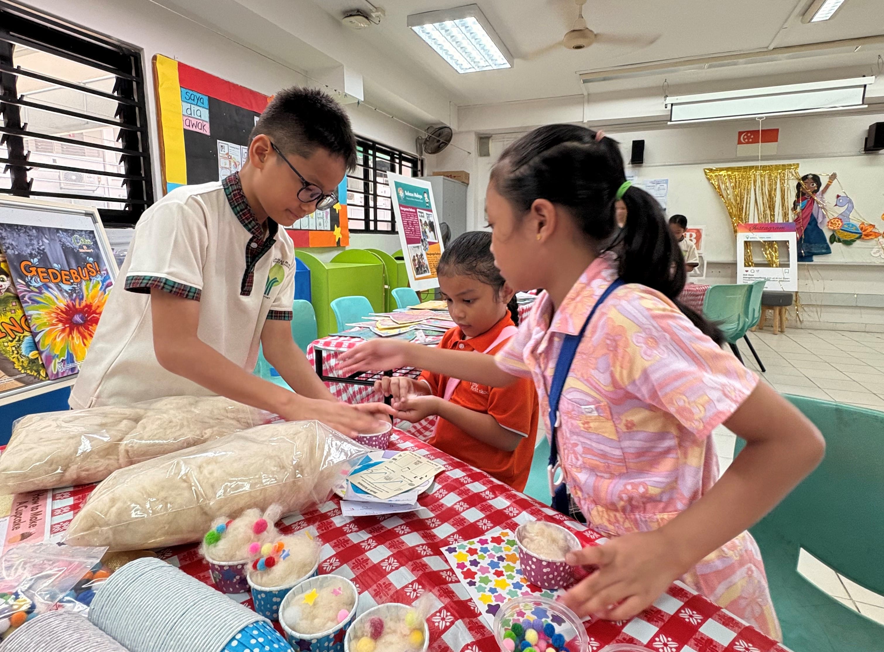 Three children at a red checkered table with craft cups, pom-poms, and cotton at a school activity fair.