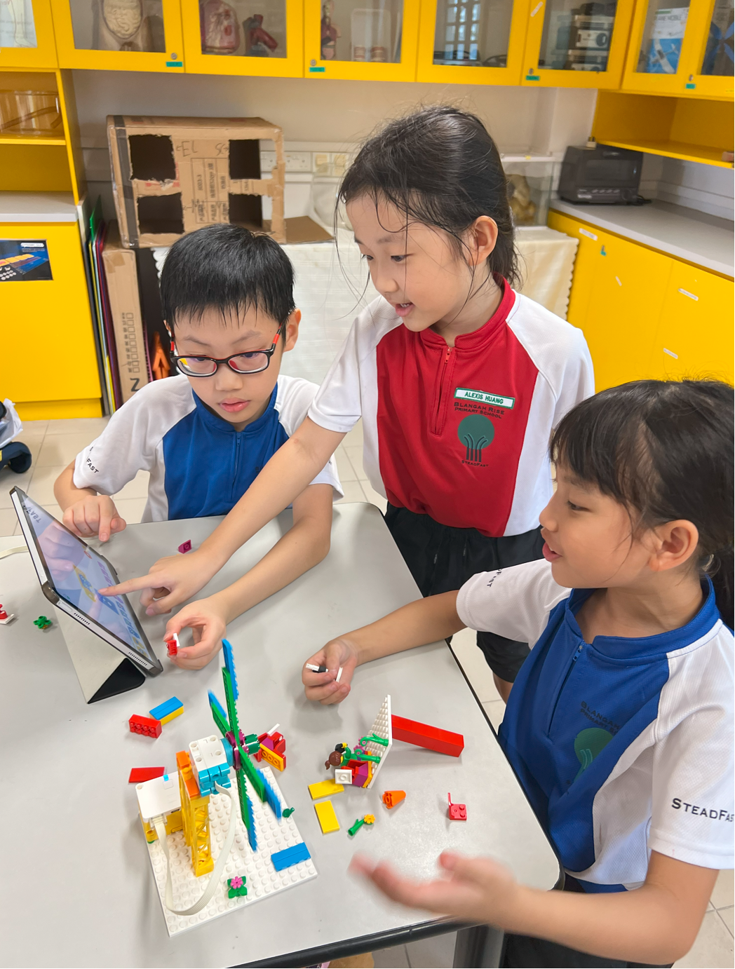 Three children in school uniforms collaborate around a colorful LEGO build and a tablet on a classroom table.