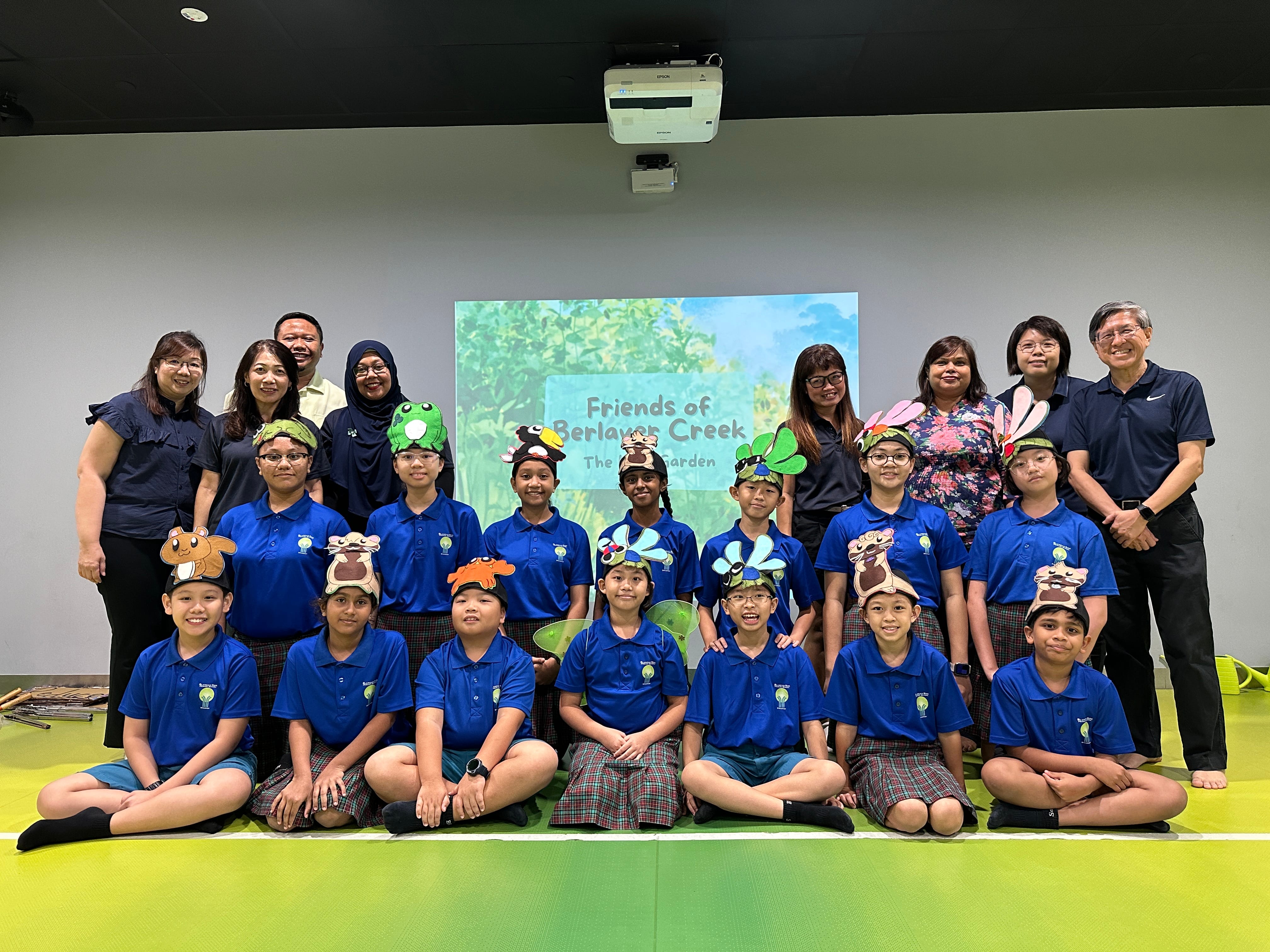Group of students in blue uniforms wearing animal headbands pose with adults before a Friends of Berlayar Creek presentation screen.