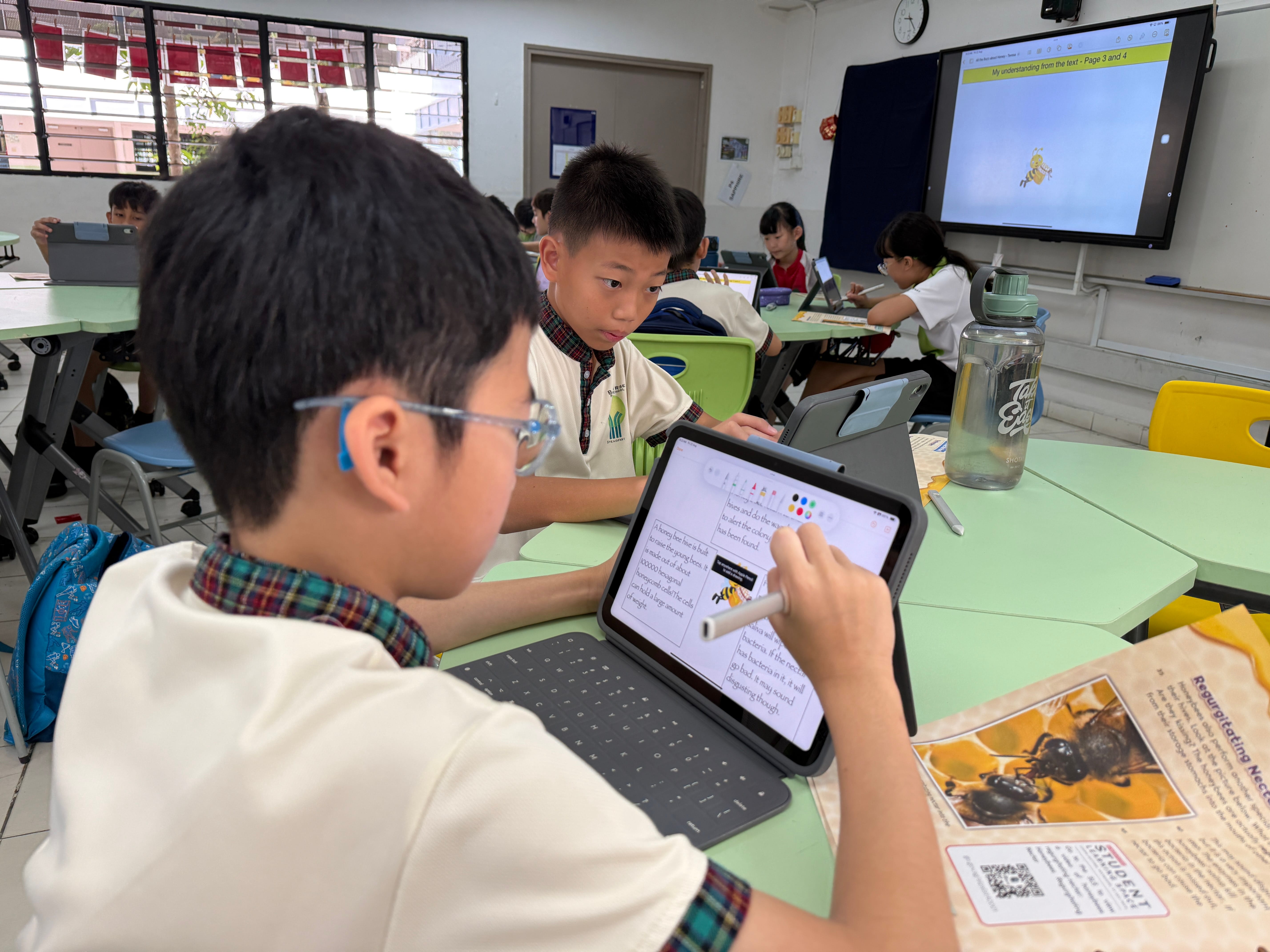 Student uses a stylus on a tablet at a classroom table while other students work on tablets nearby.