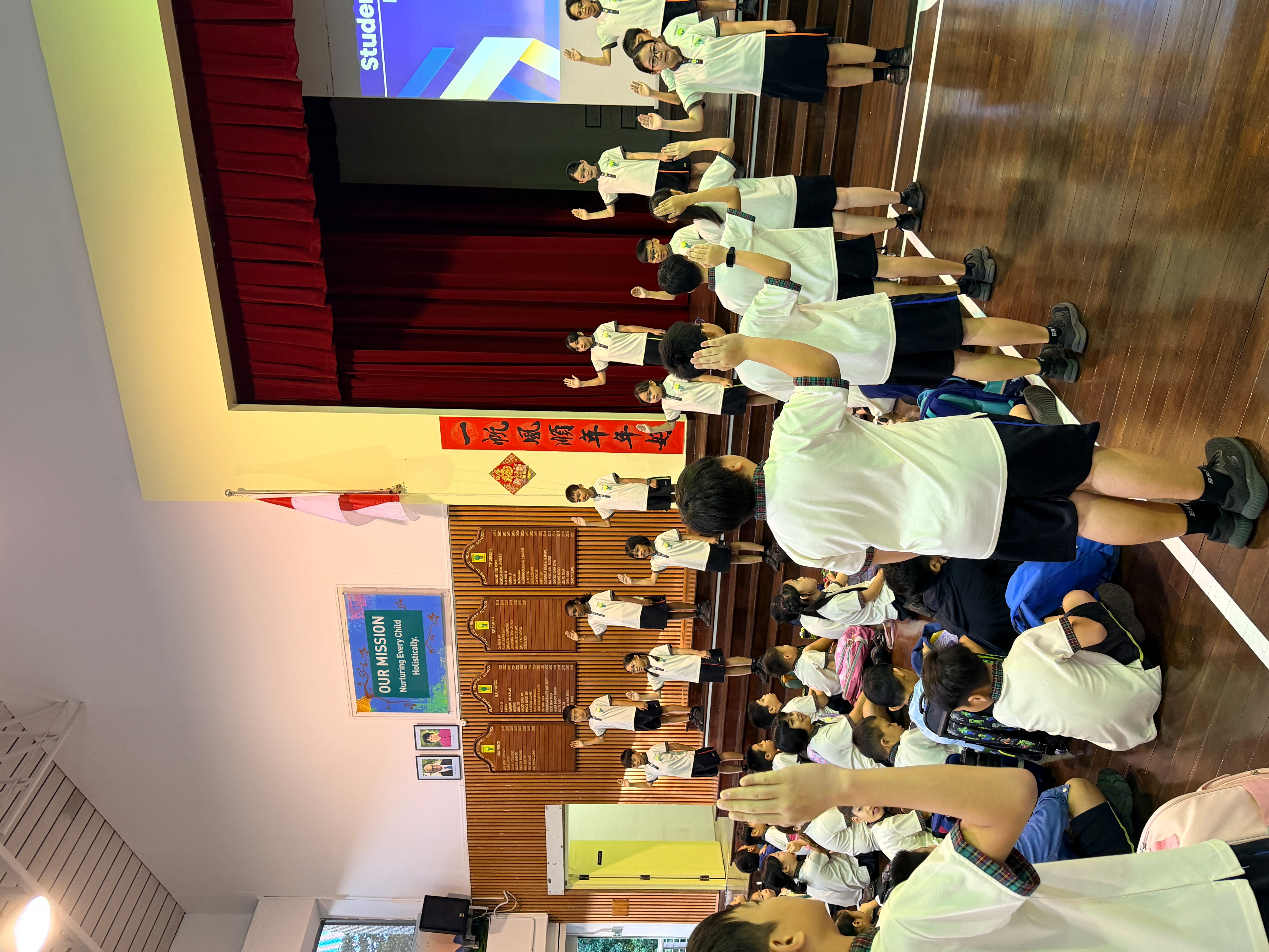Students in white uniforms in a school hall, many raising hands toward a stage with red curtains.