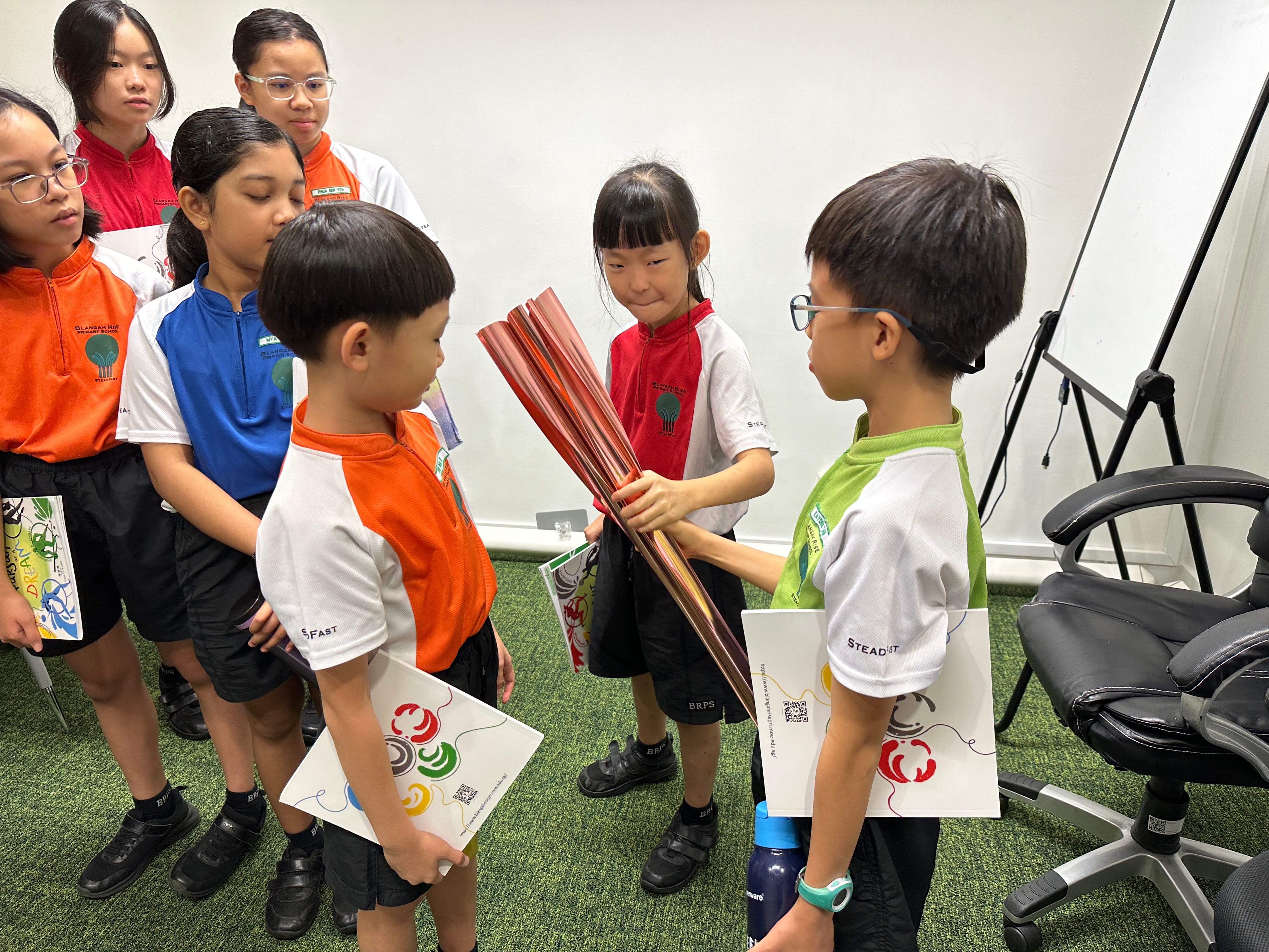 A group of young students in school uniforms interact indoors, with two boys facing each other holding red sheets.