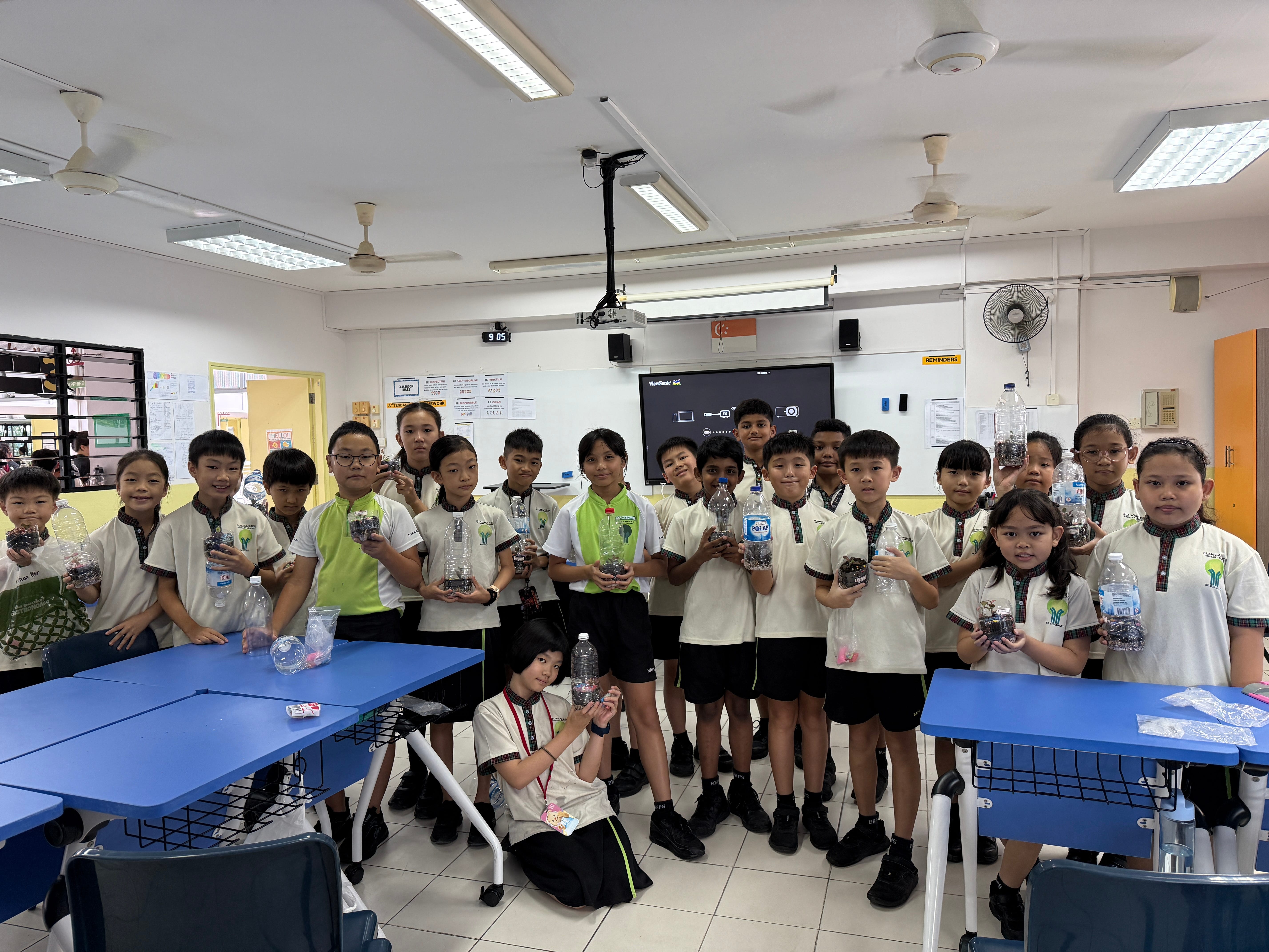 Group of schoolchildren in a classroom holding plastic bottles filled with small items, standing by blue tables.