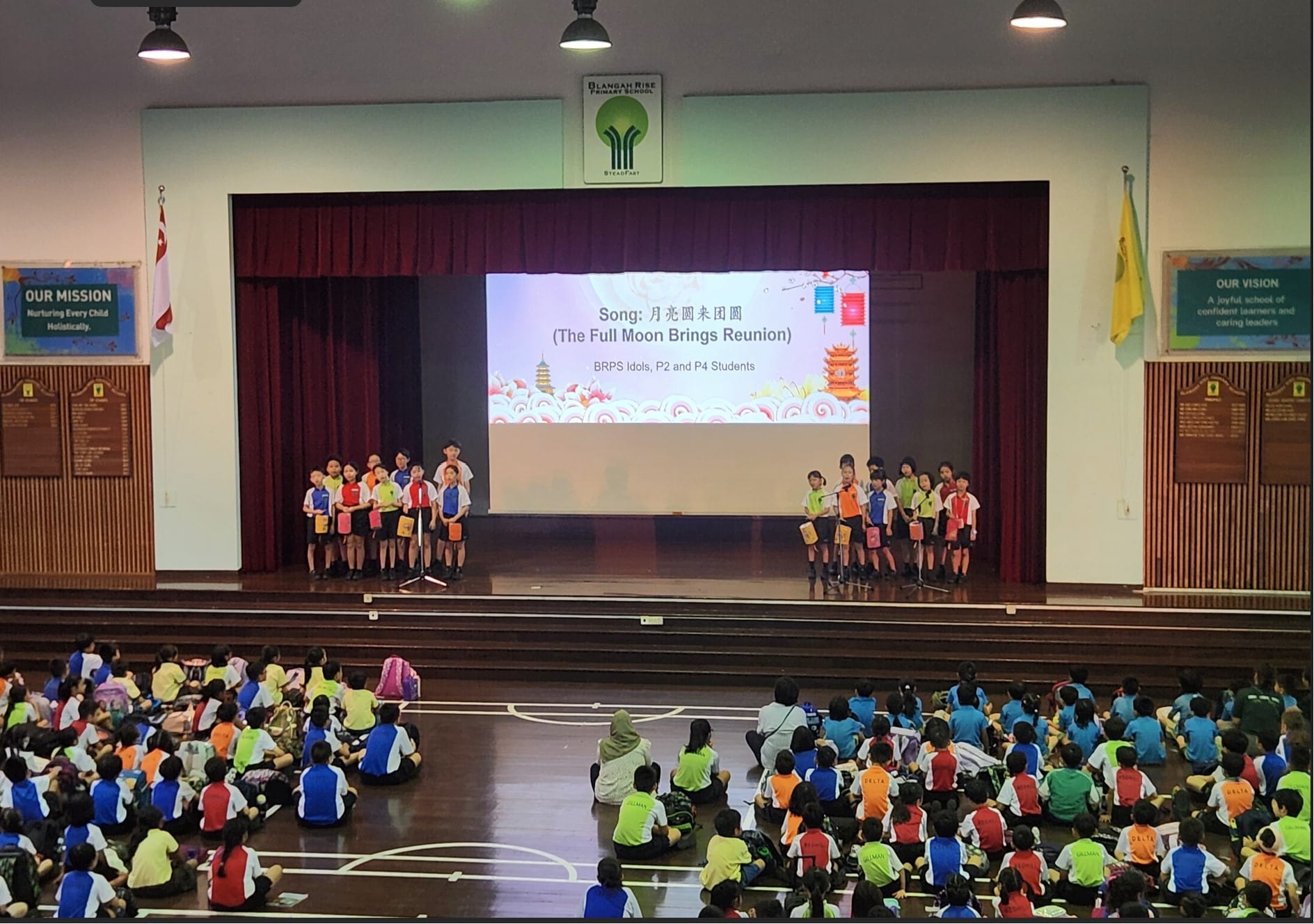 Students perform on a school stage holding lanterns while a large seated audience watches a Full Moon Reunion song presentation.