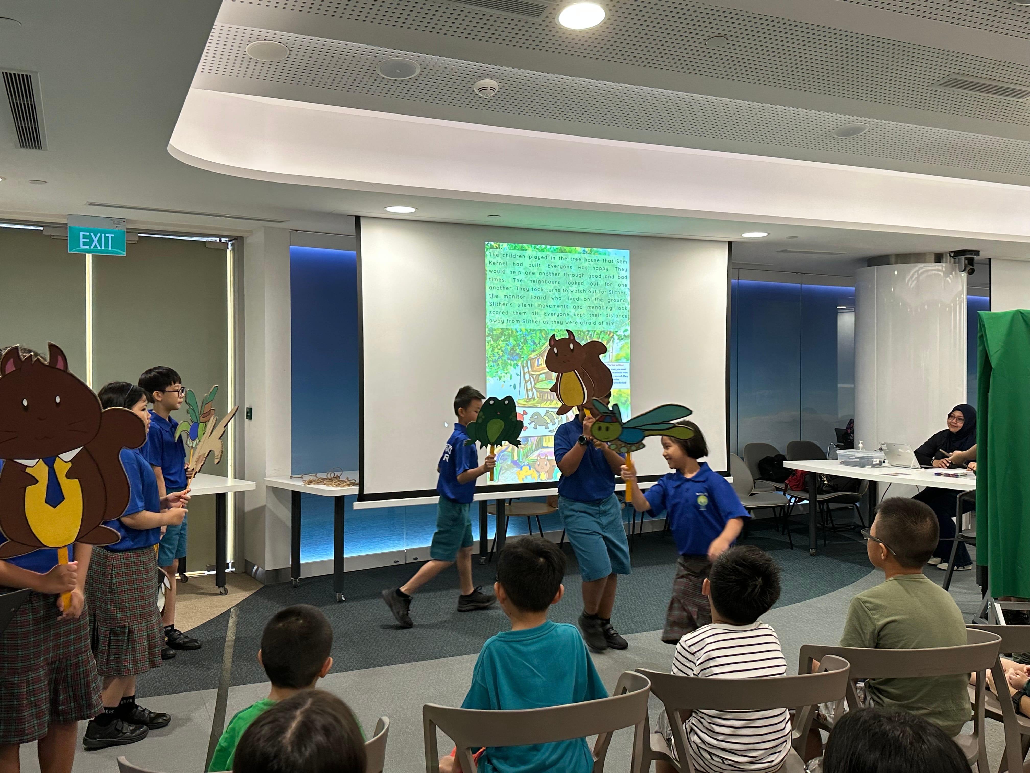 Children in blue uniforms hold animal puppets during a storytime performance in front of a projected storybook slide.