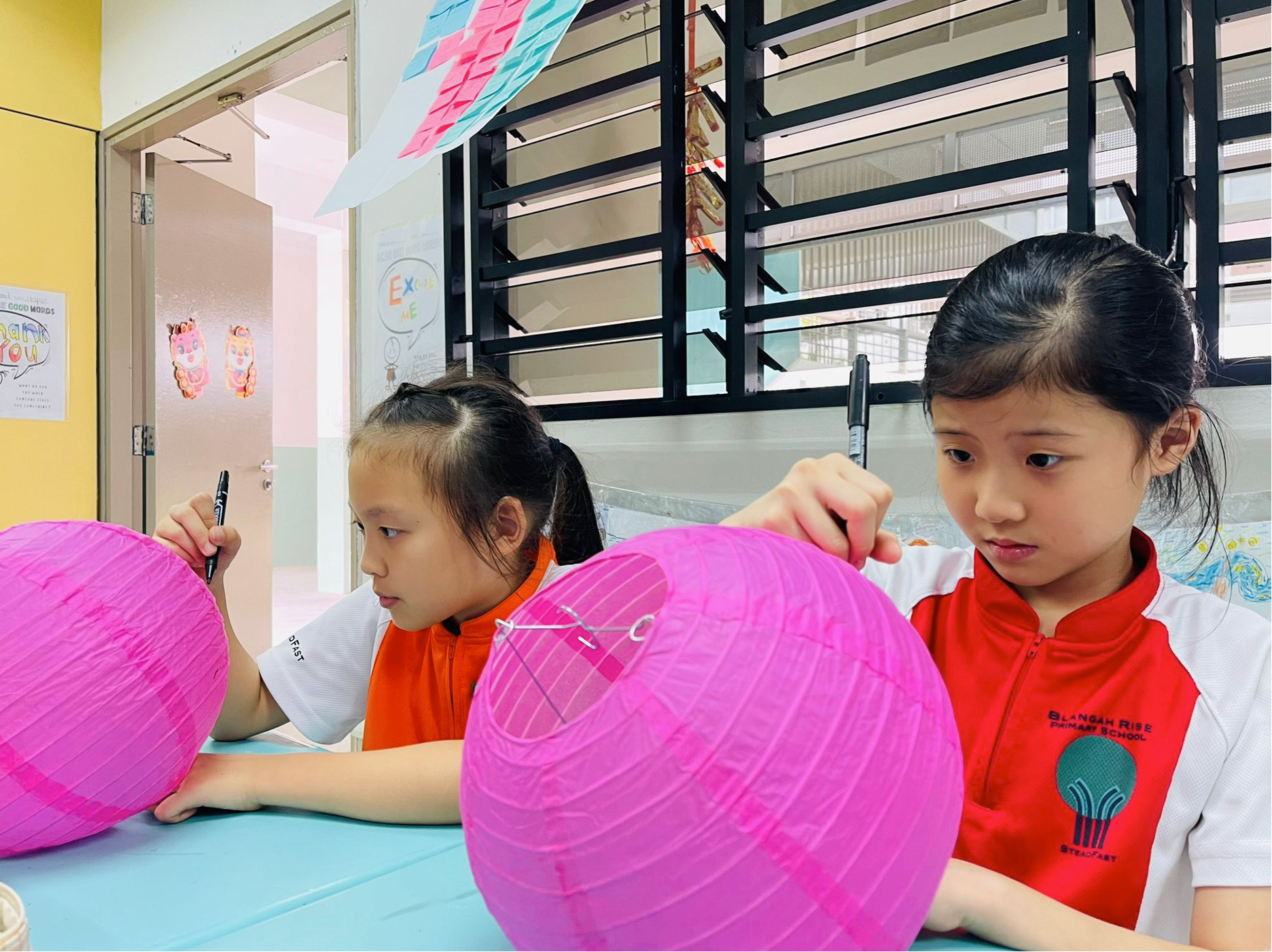Two girls in red-and-white school uniforms draw on large pink paper lanterns at a table in a classroom.