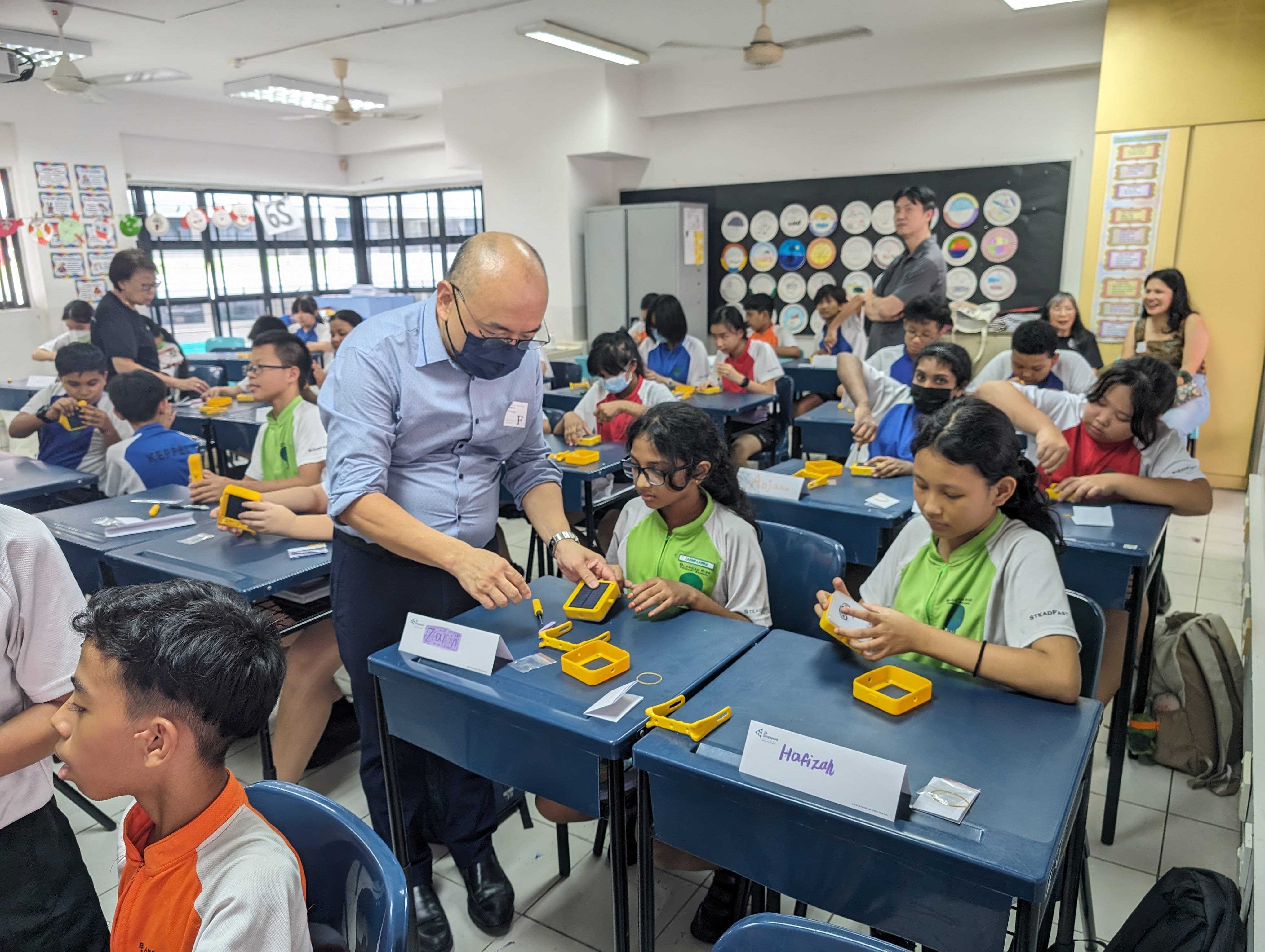 A masked adult man leans over to assist students at desks with yellow trays in a busy classroom.