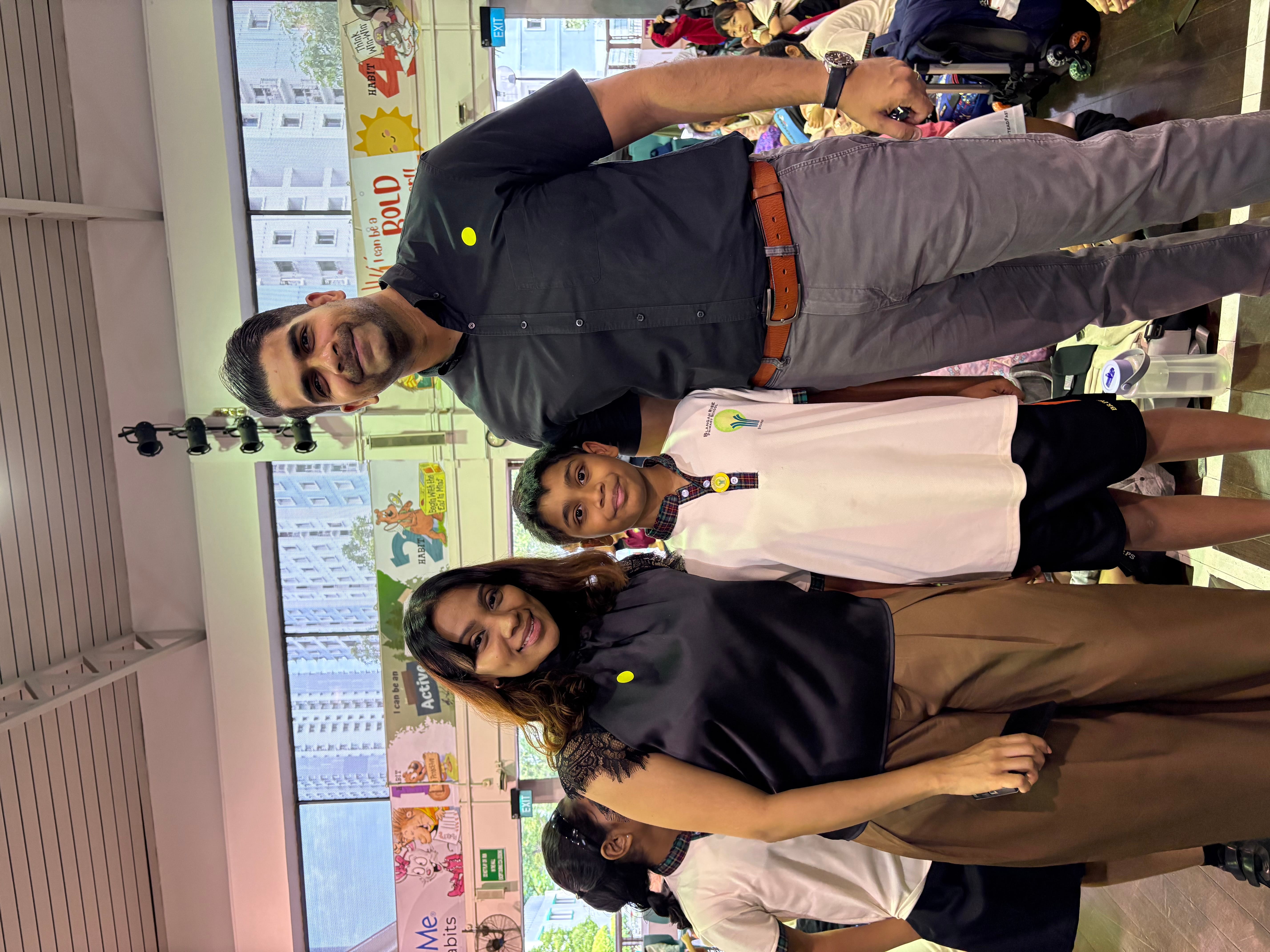 Man and woman stand with a schoolboy in a white uniform at an indoor event with people seated behind.