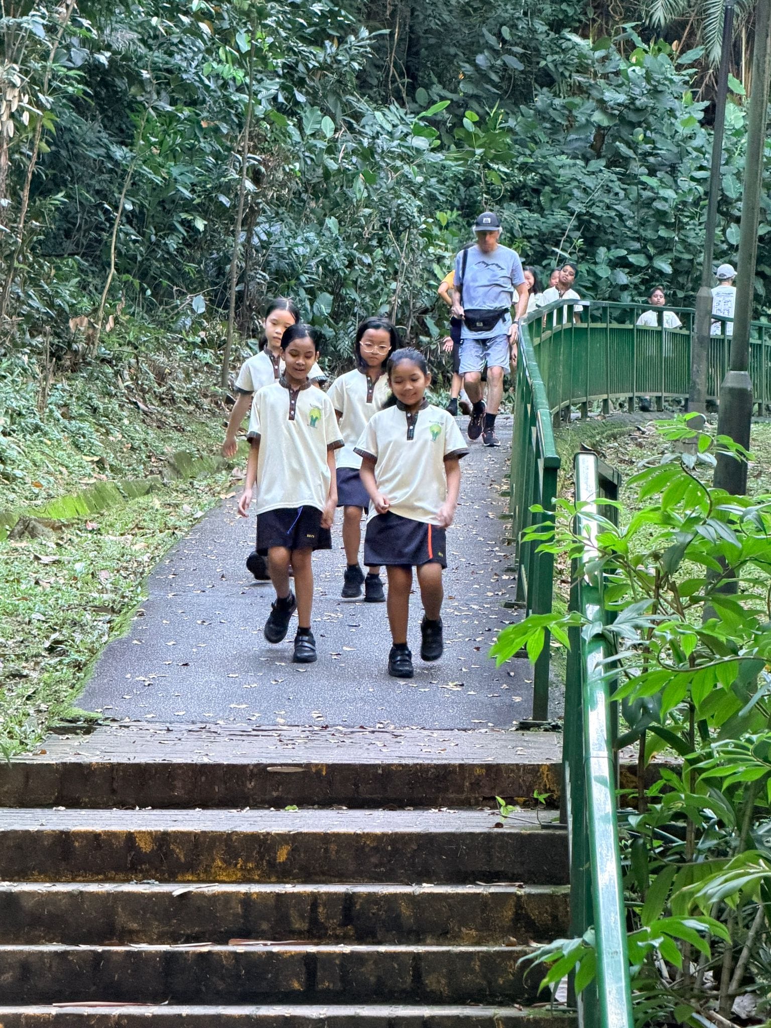 Children in white uniforms walk down a forested park path with green railings, followed by an adult in gray.