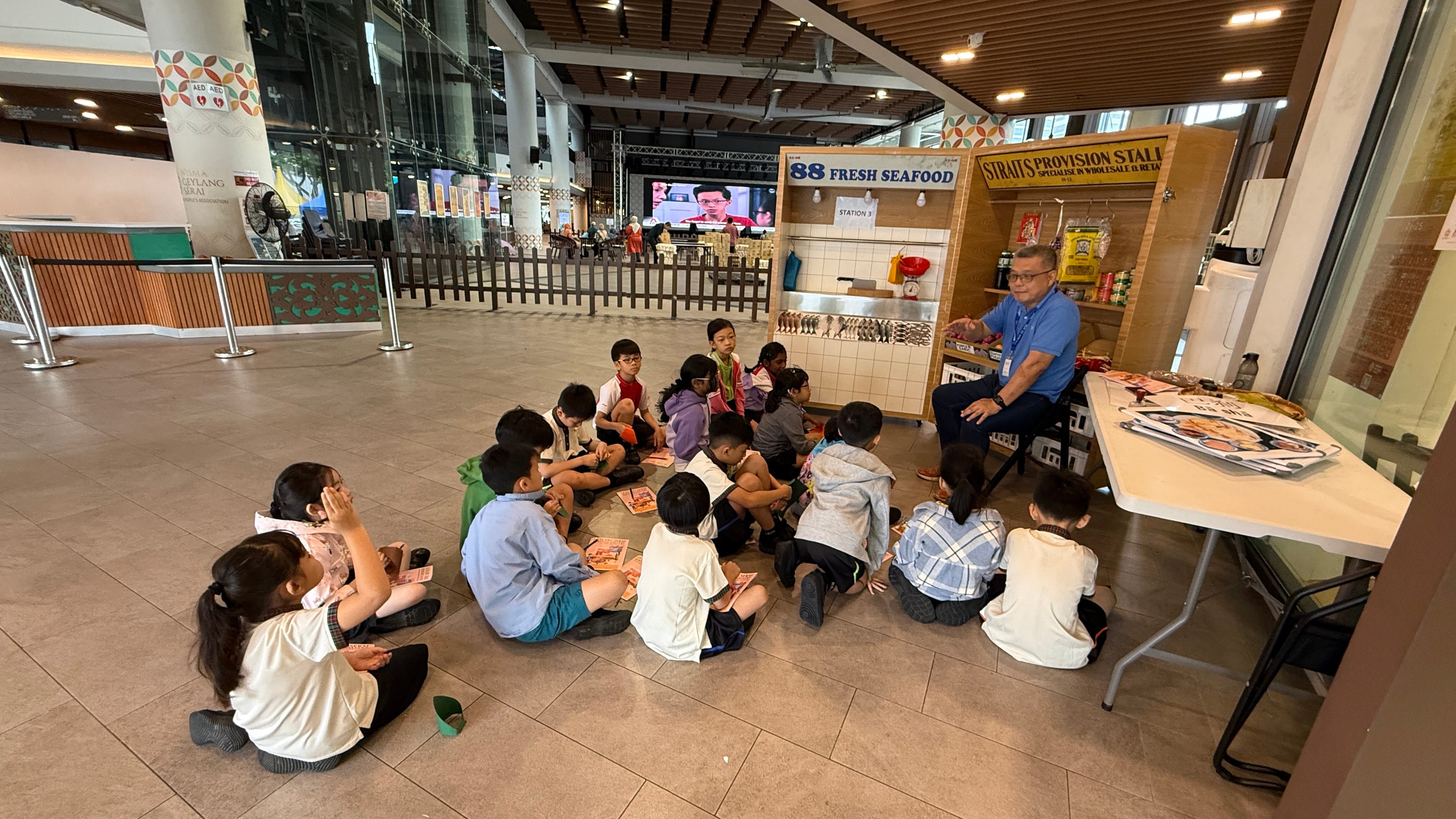 A man seated at a table teaches a group of young children sitting on the floor near market stall displays indoors.