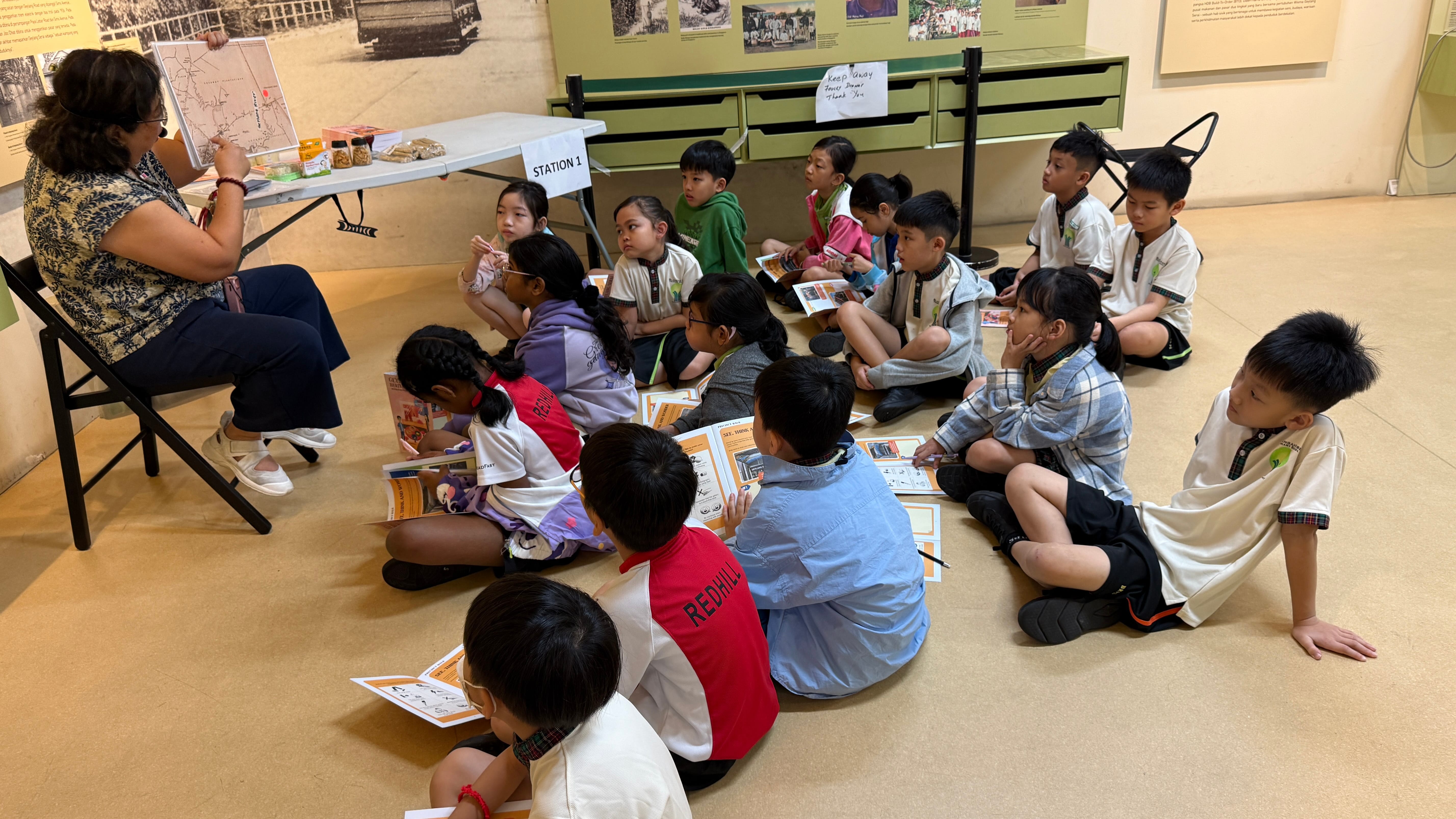 A teacher sits on a chair facing a group of young children seated on the floor at a museum Station 1 activity.