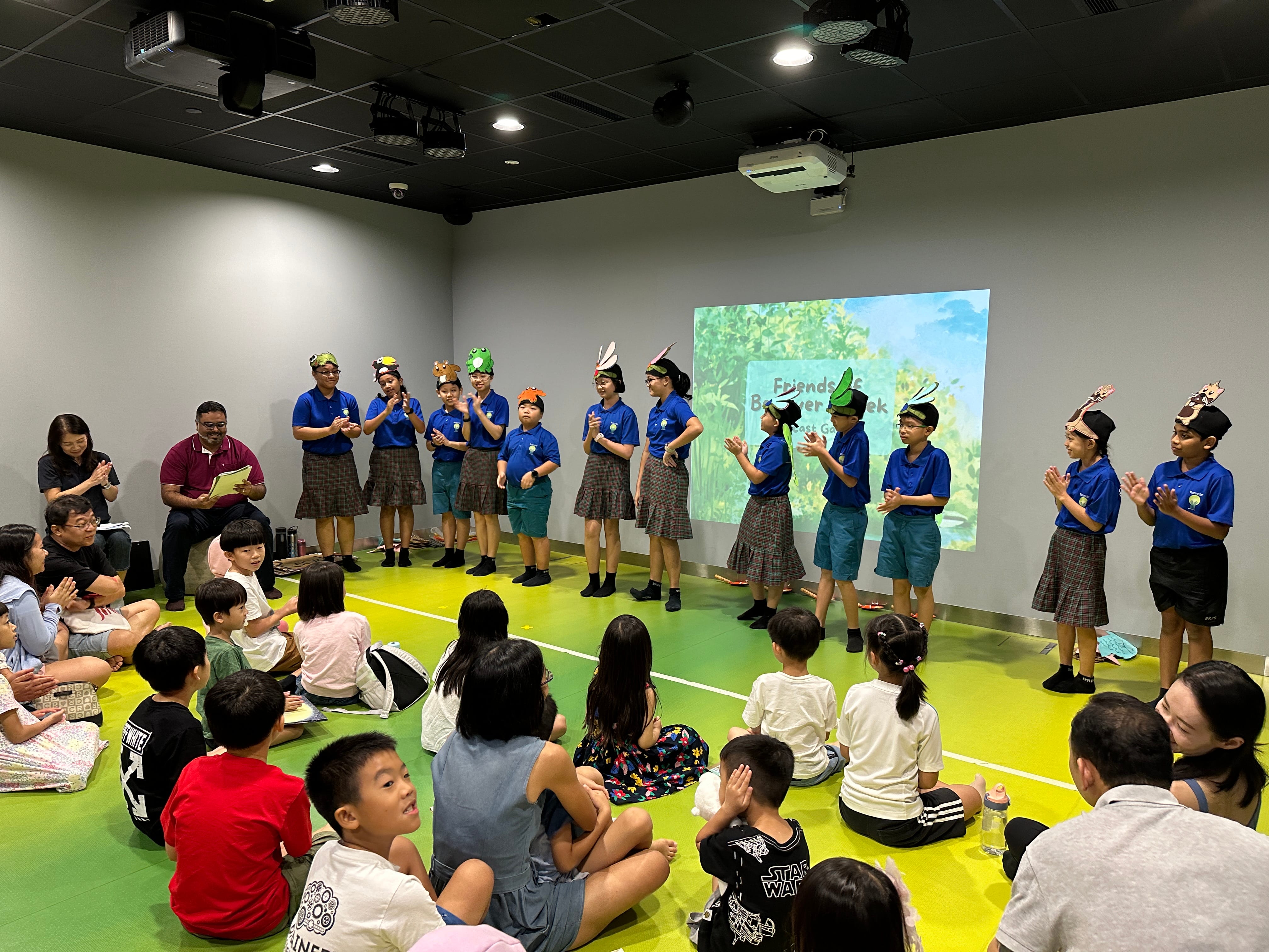 Children in school uniforms on a stage clap as seated kids watch; projector shows forest scene behind.