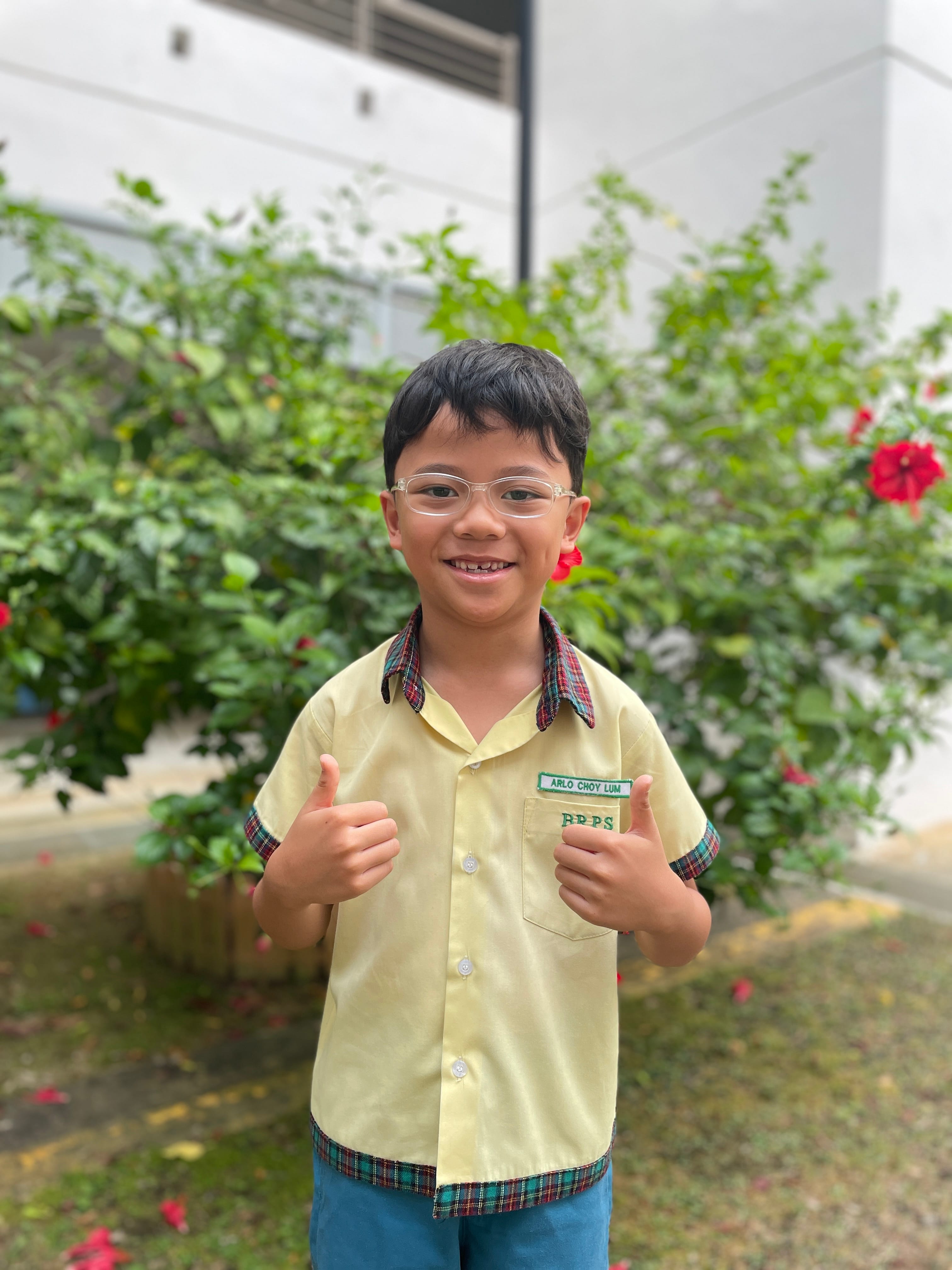 A smiling young boy in a yellow school uniform with glasses gives two thumbs up in front of green bushes with red flowers.