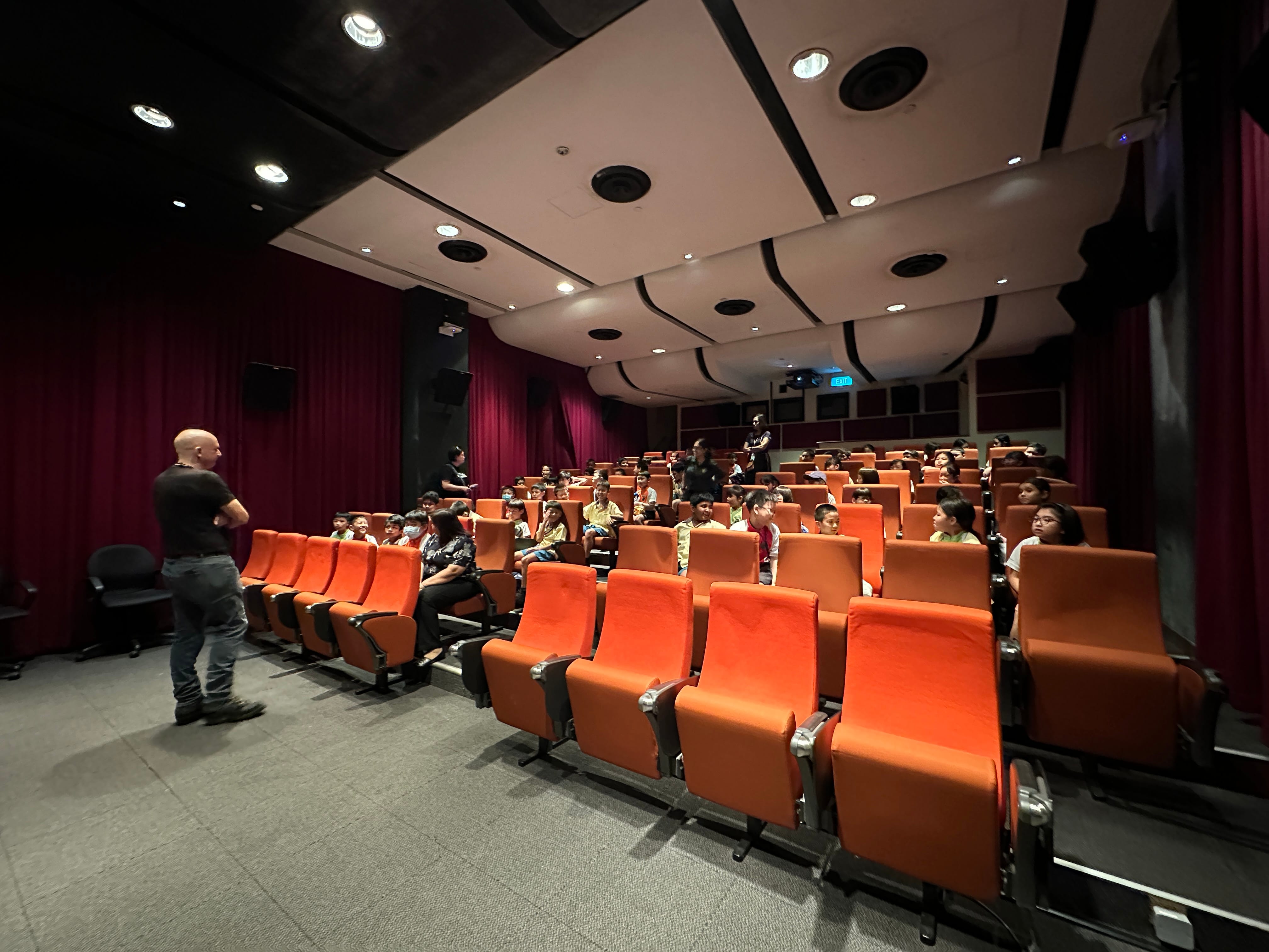 A man stands at the front of a small theater with orange seats, addressing a group of children seated in rows.