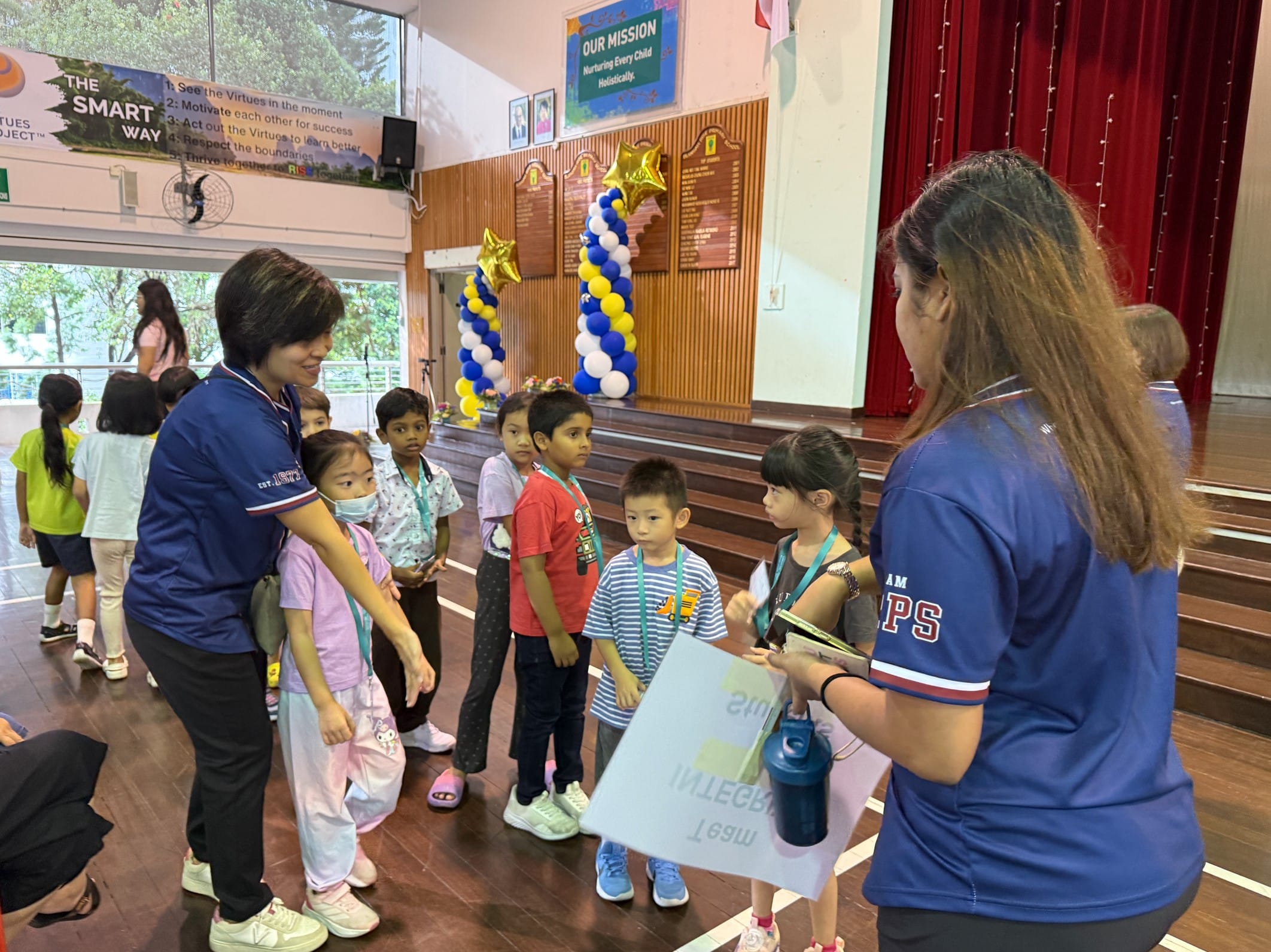 Two adults in blue school shirts guide a group of young children in a decorated school hall with balloon columns.