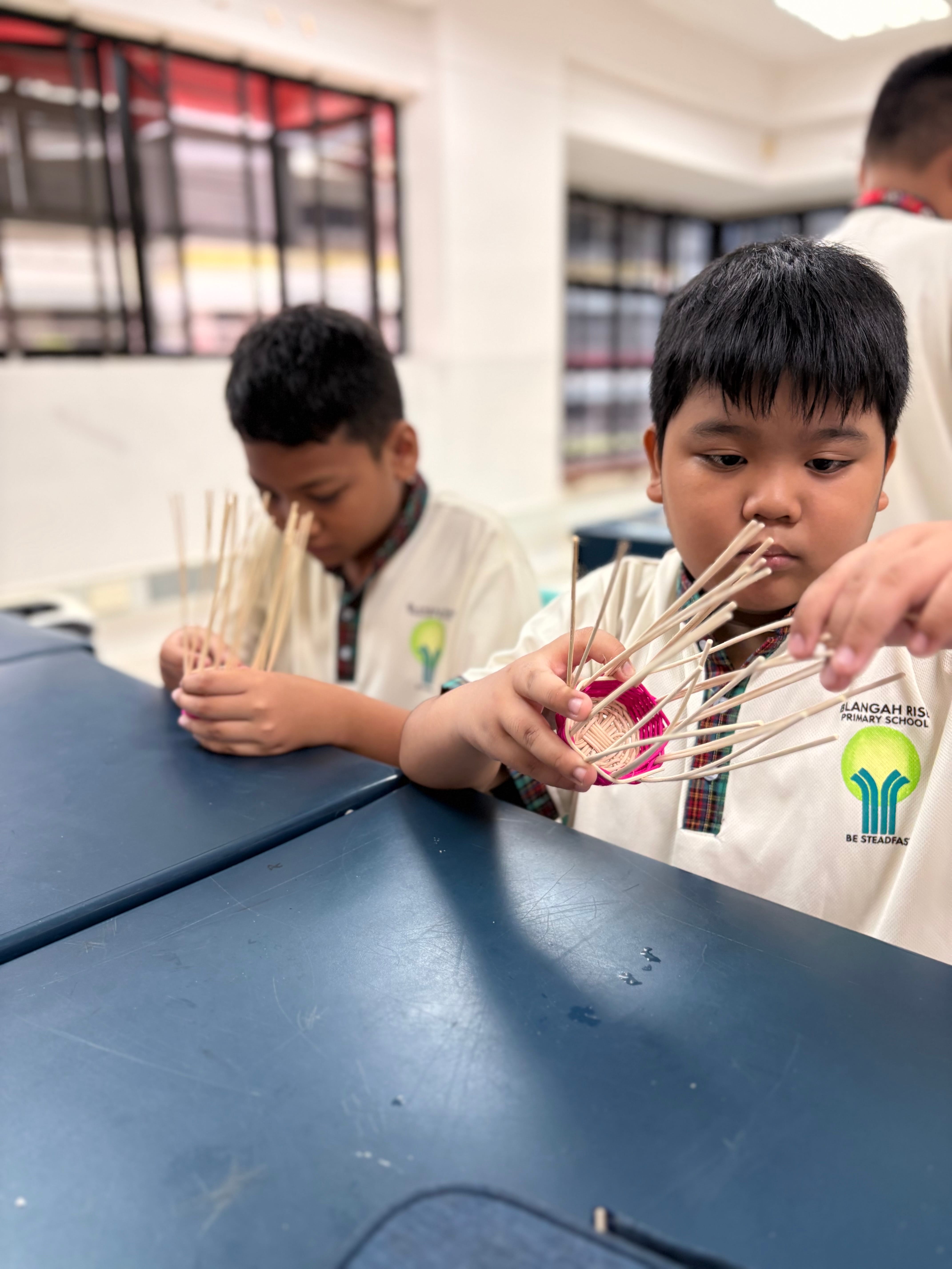 Two primary school boys in white uniforms weave thin bamboo sticks into small baskets at a blue desk.