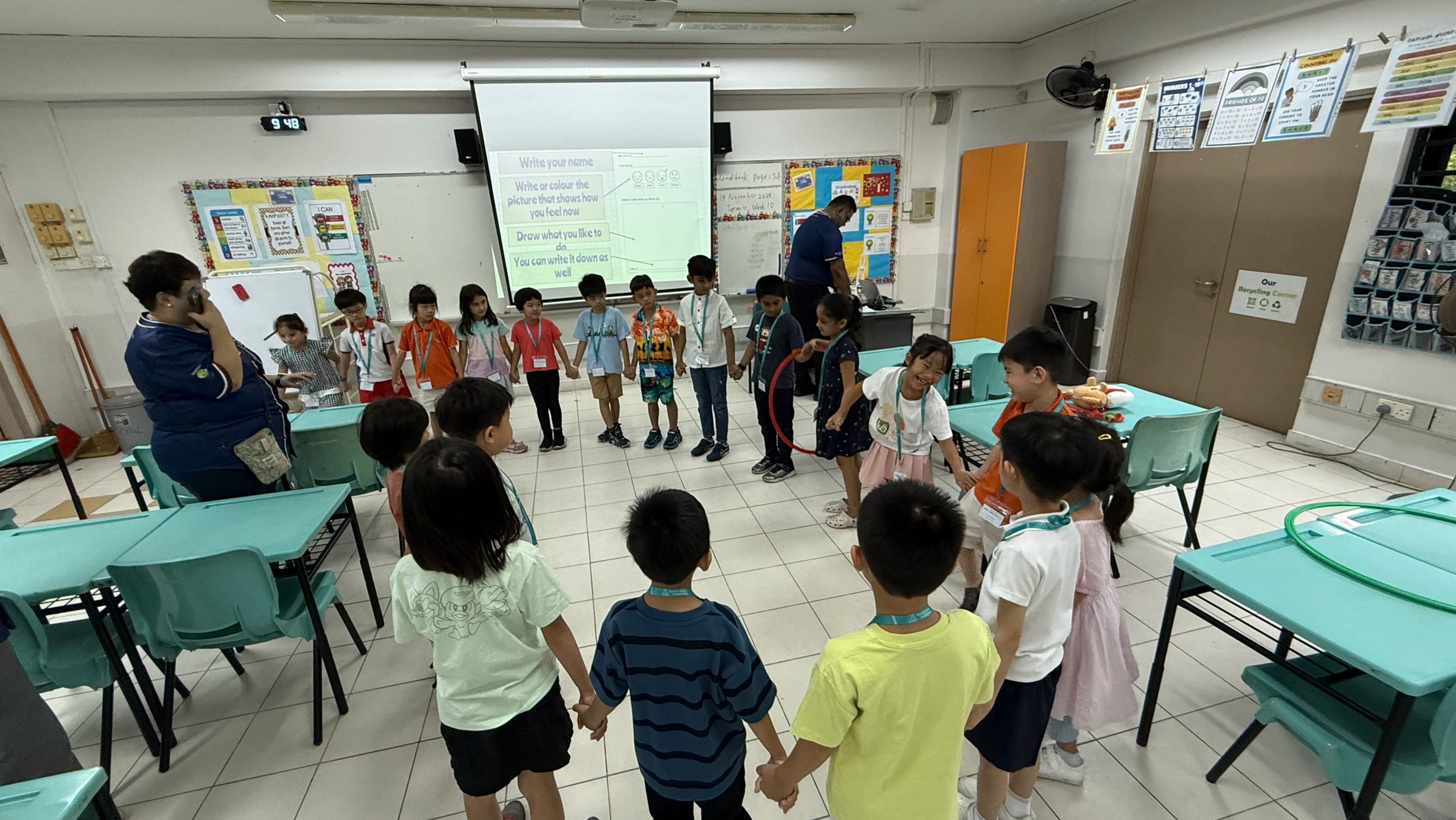 Young children hold hands in a circle in a colorful classroom with a projector screen displaying a writing activity.